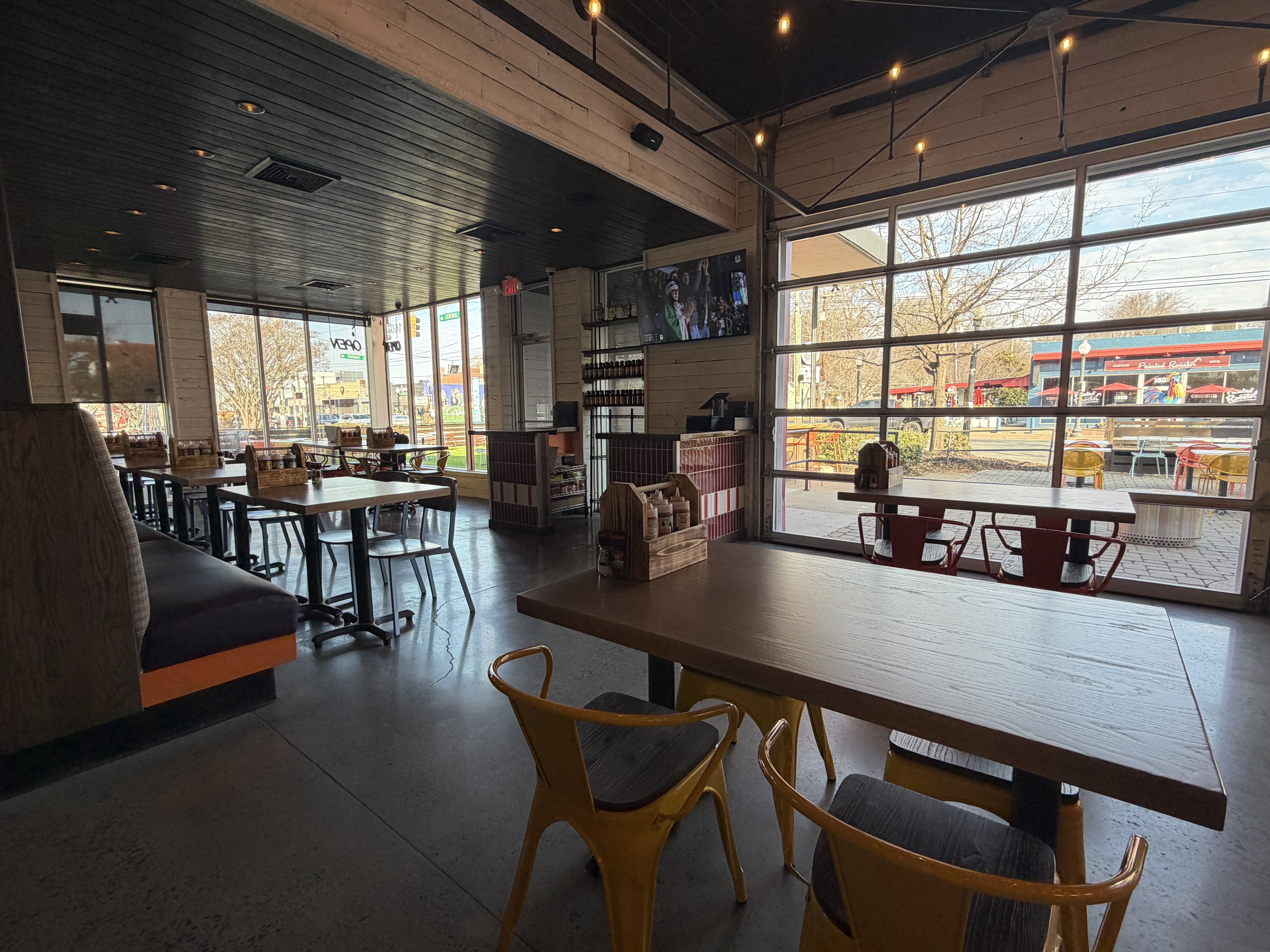 Empty casual dining area with wooden tables, yellow metal chairs, a TV on the wall, and large windows showing outdoor seating and a street scene with trees and buildings.