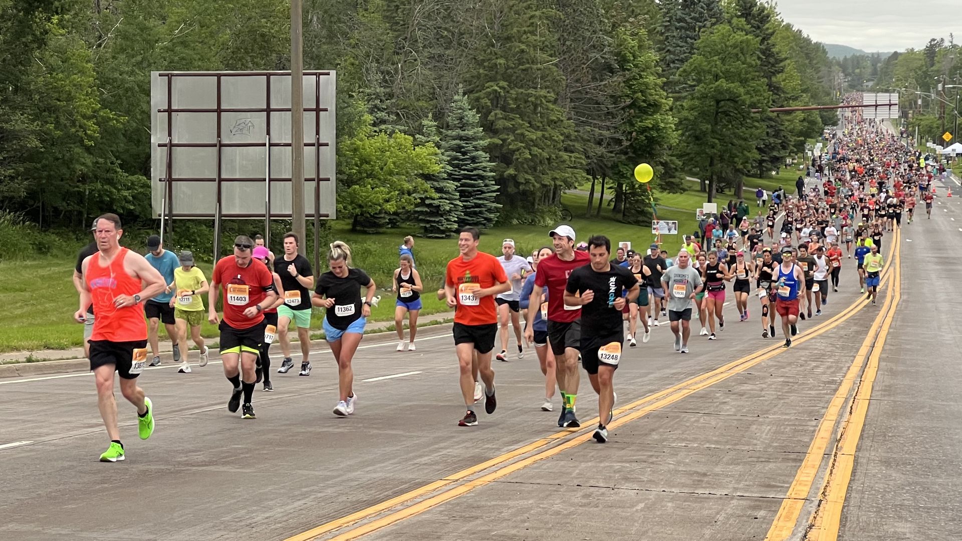 Thousands of runners run up a closed street toward the photographer, who’s capturing the image looking downhill. Green trees surround the scene.