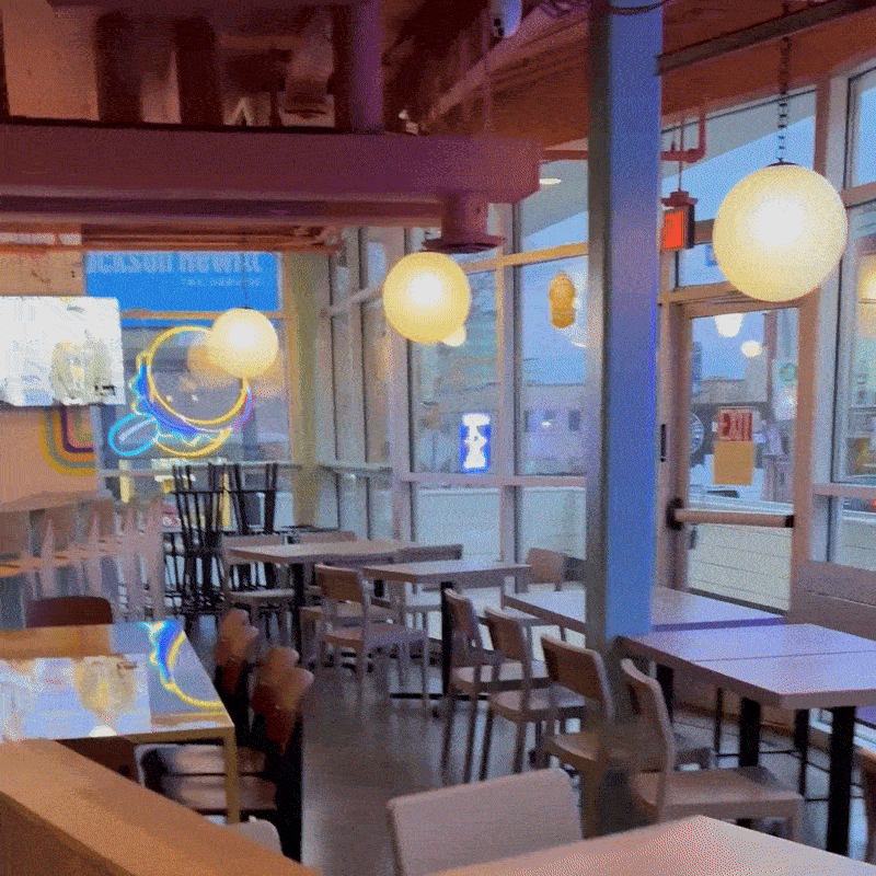 A panning shot of the inside of a restaurant showing tables, a bar, and neon blue and yellow lights. 