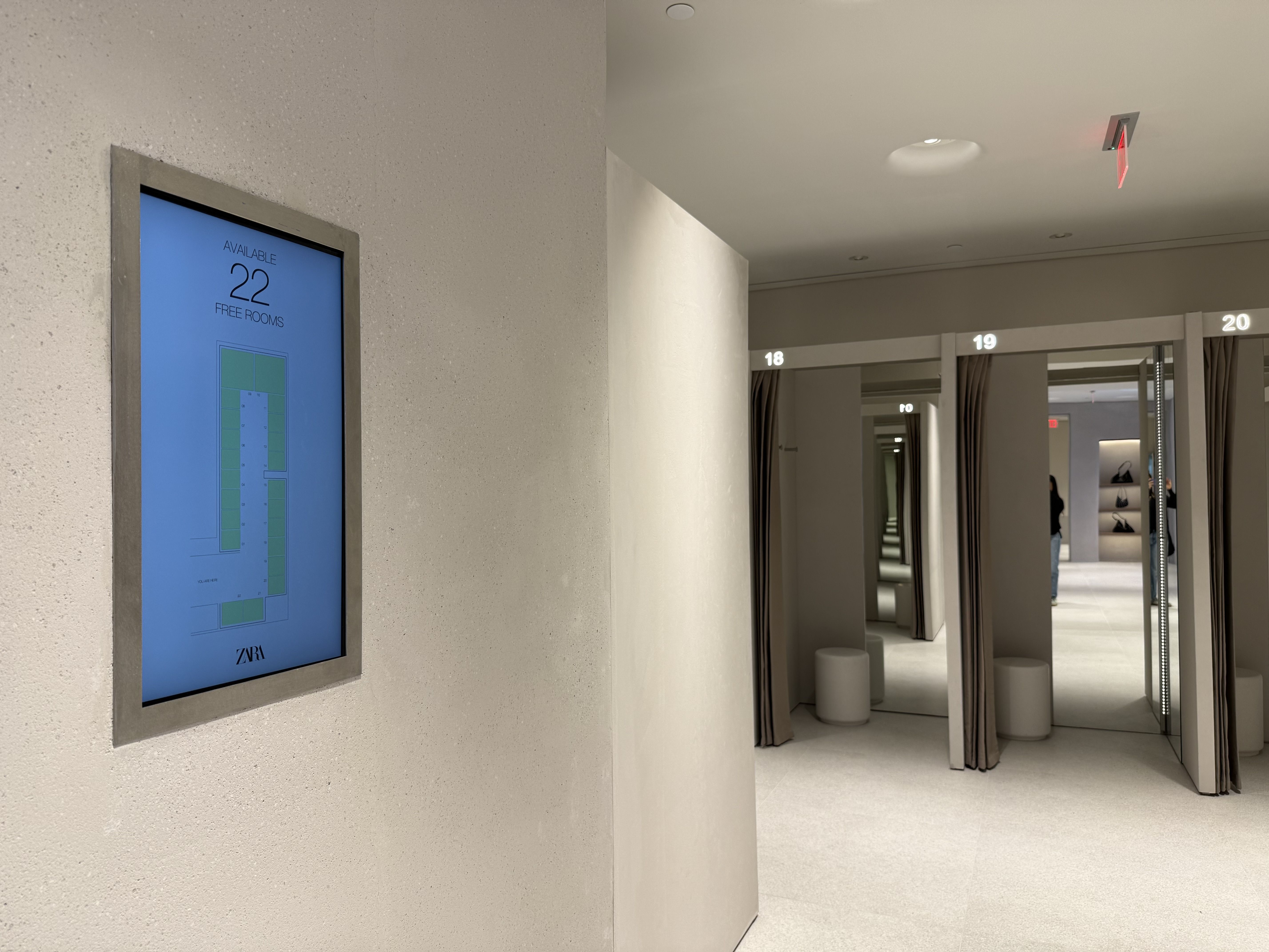 View of a modern fitting room area with numbered stalls 18, 19, and 20 and a screen on the wall showing 22 free rooms available at Zara store.