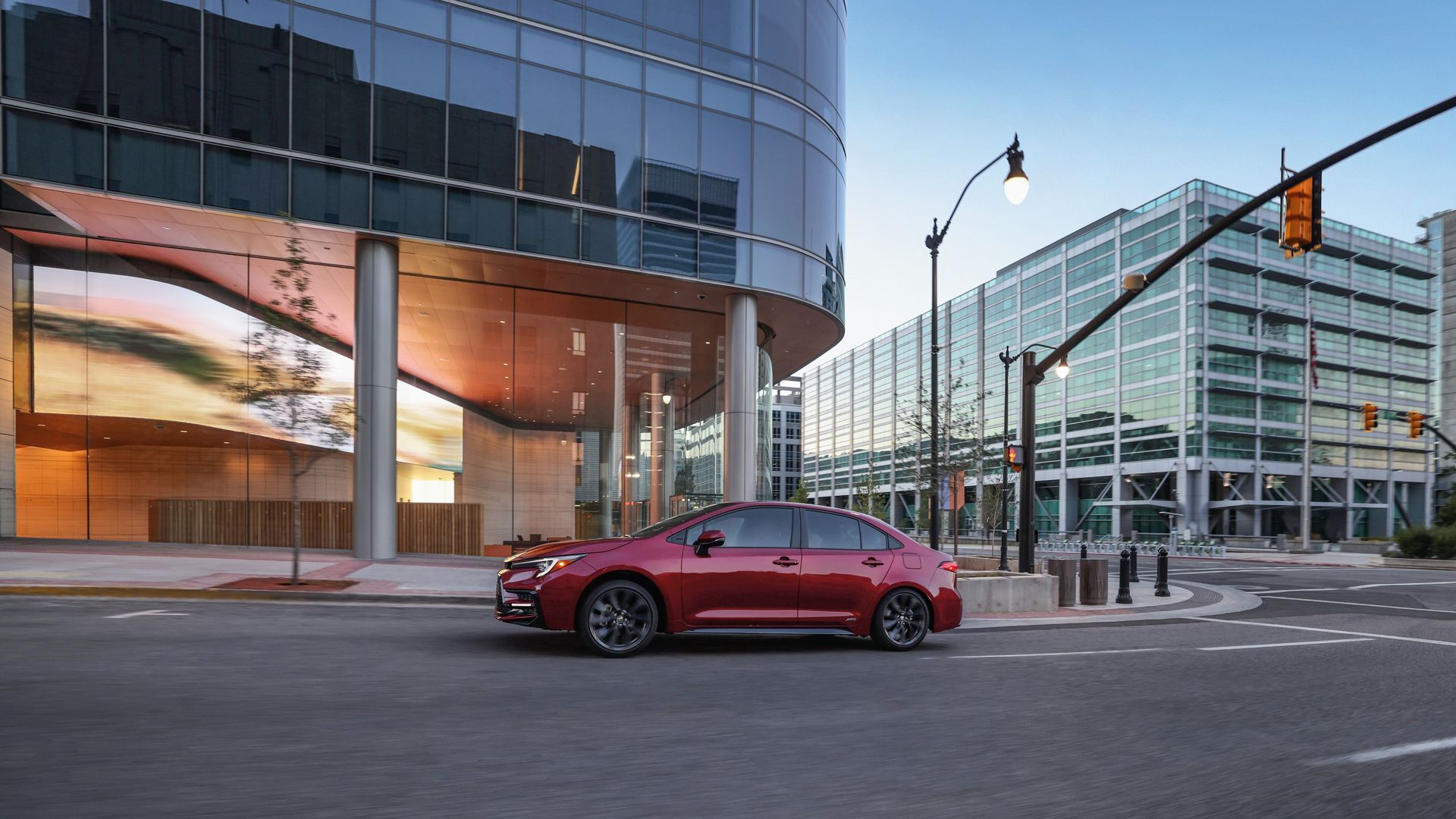Red Toyota Corolla hybrid sedan car parked on a city street near modern glass buildings with streetlights and traffic signals visible.