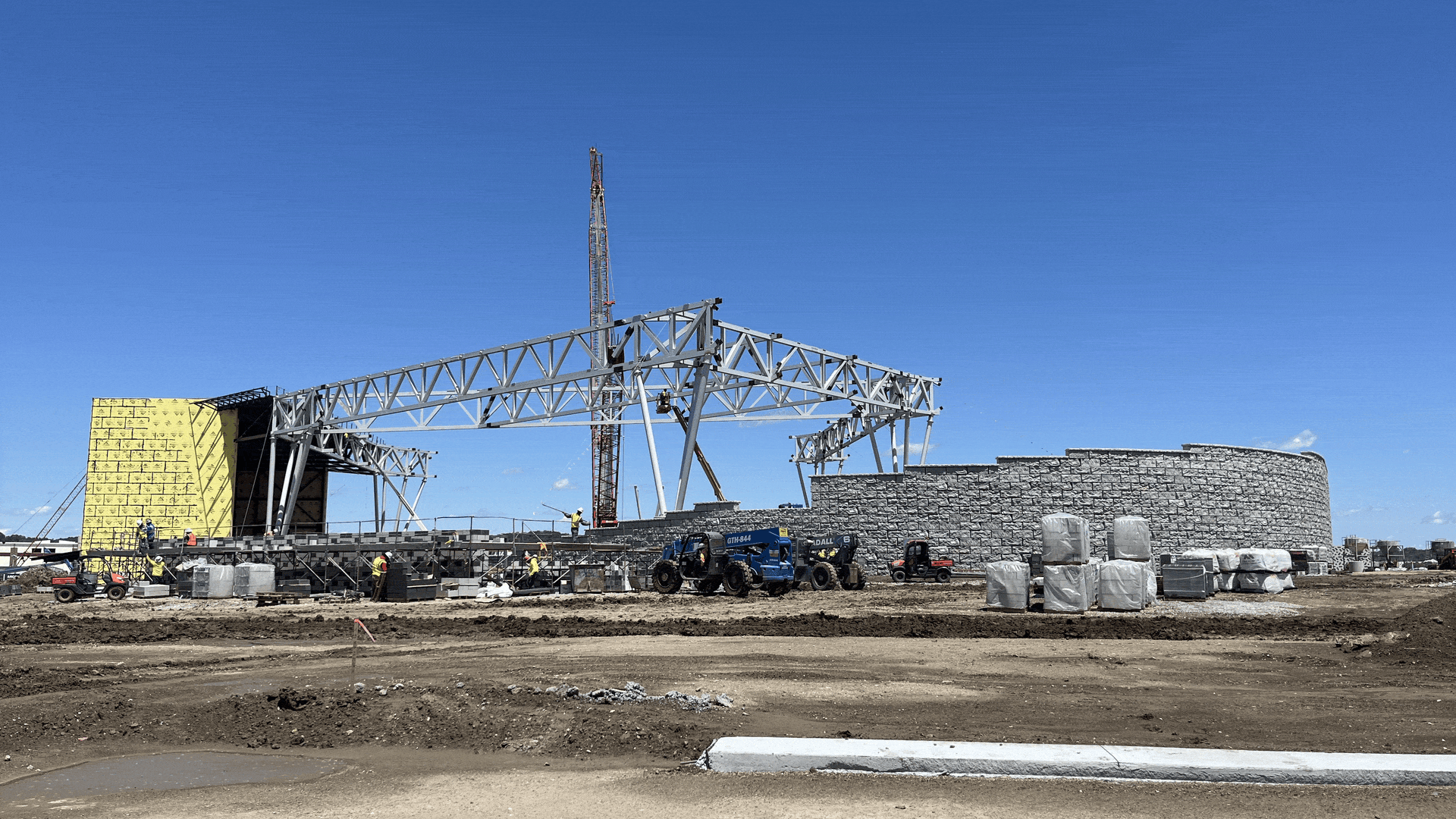 Construction site with large gray metal framework and a crane in the background. Yellow insulated wall on the left, stone wall on the right, workers and machinery on dirt ground under a clear blue sky.