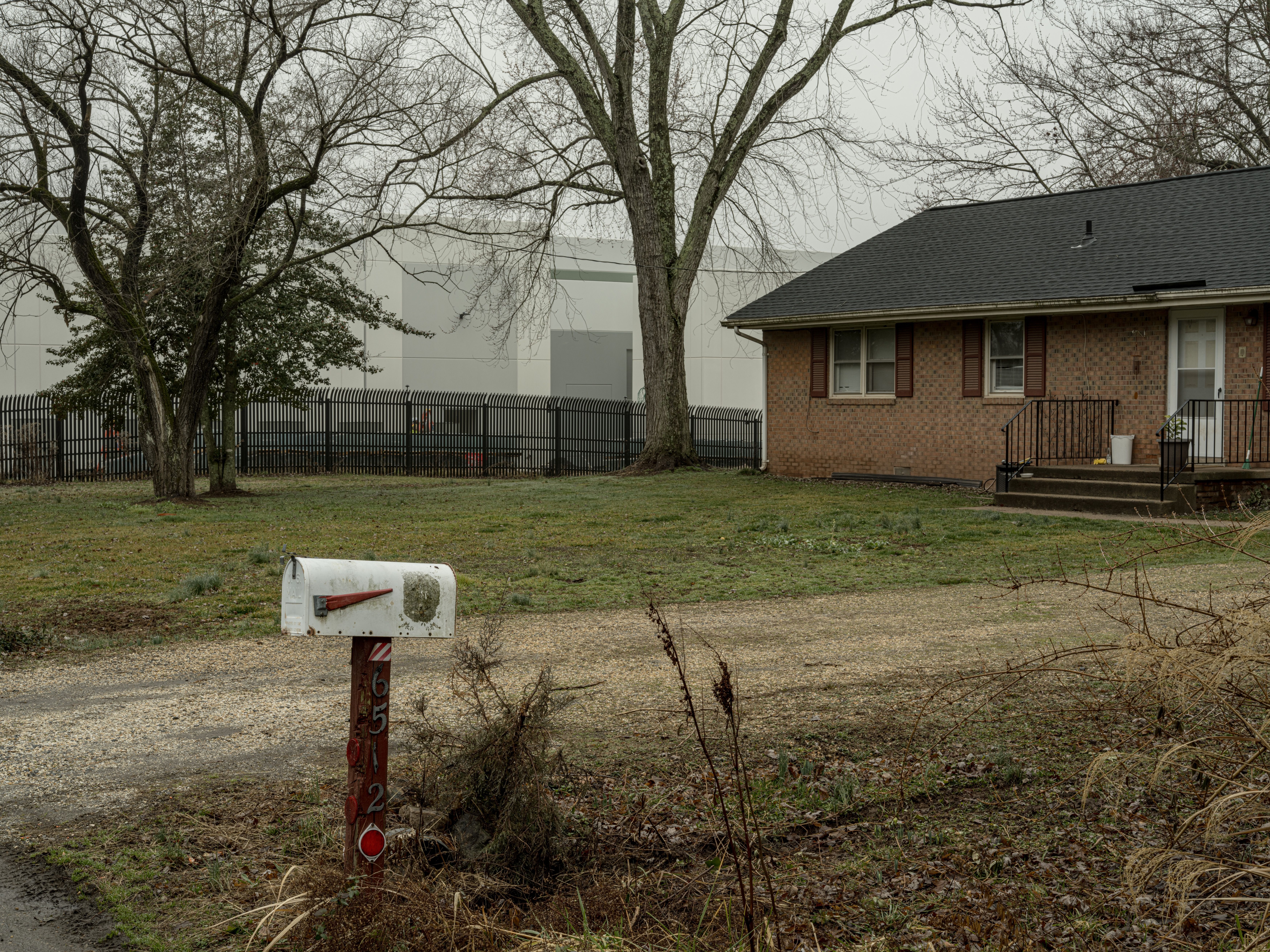 Leafless trees frame a yard with a brick house on the right and a white modern building in the background. A black metal fence runs across the scene; a white mailbox on a wooden post sits in the foreground.