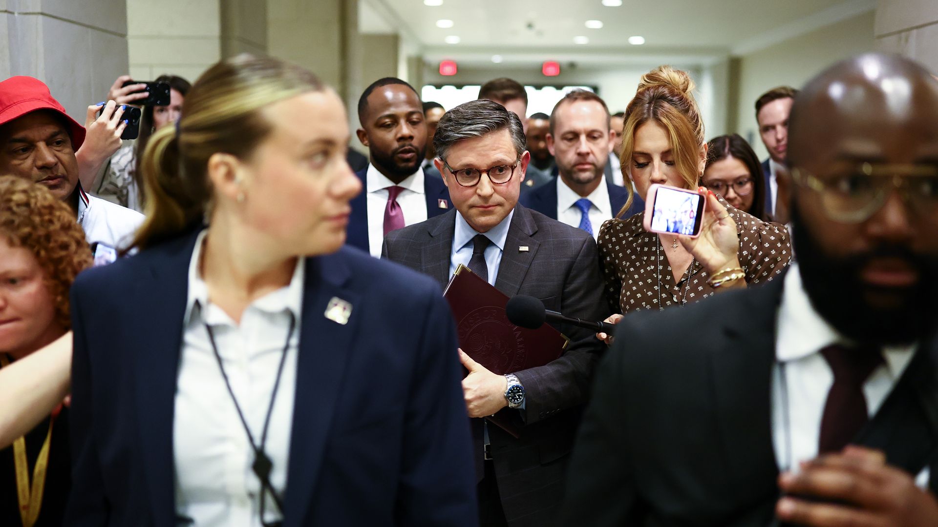 A crowded hallway of suited professionals. In the center, a man in a dark suit and glasses looks down, while others around him hold phones and record, including a woman on the right with a pink phone.