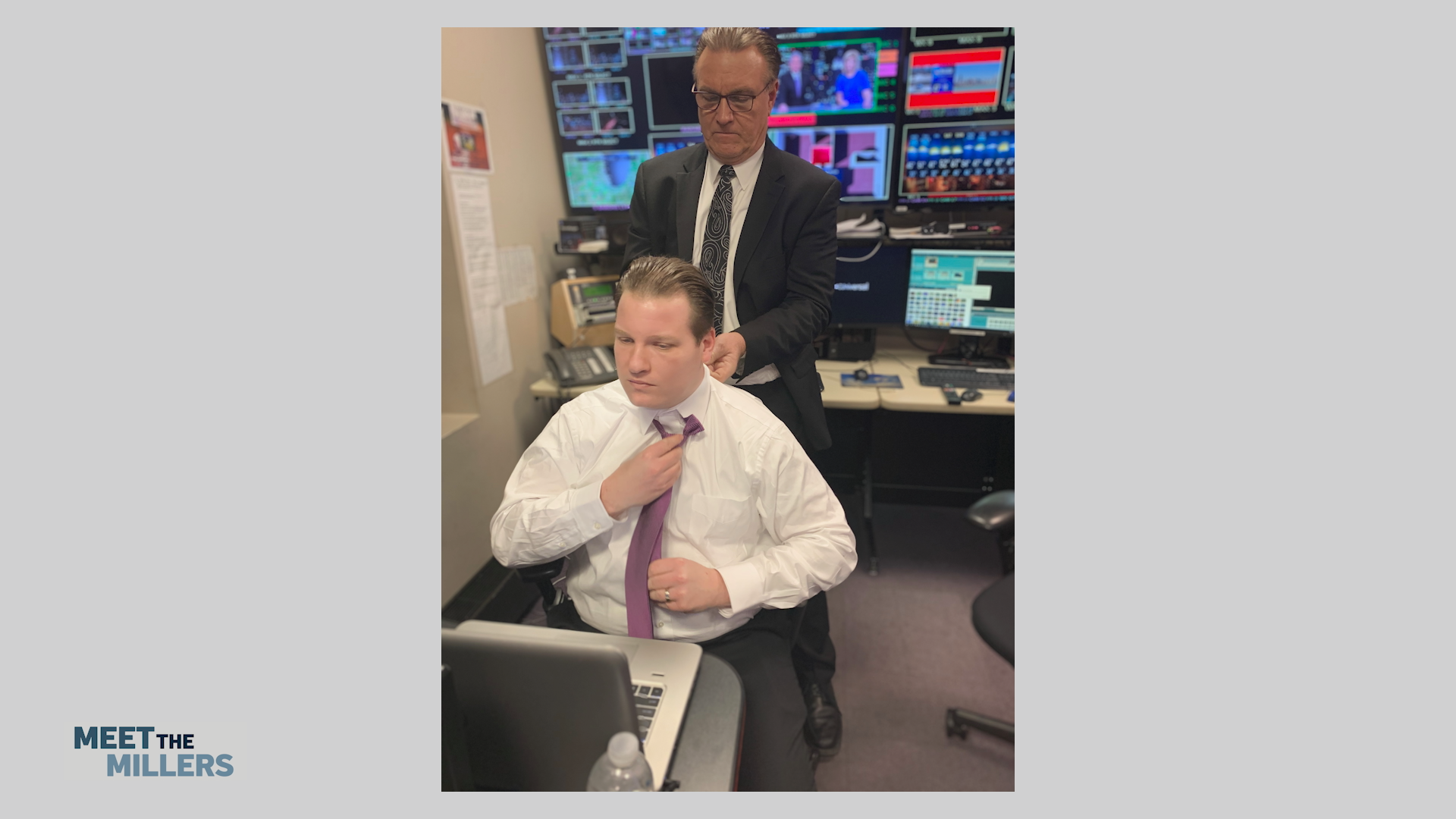 Two men in a TV control room; a seated man in a white shirt and purple tie adjusts his tie as a suited colleague stands behind him, with multiple monitors and equipment in the background.