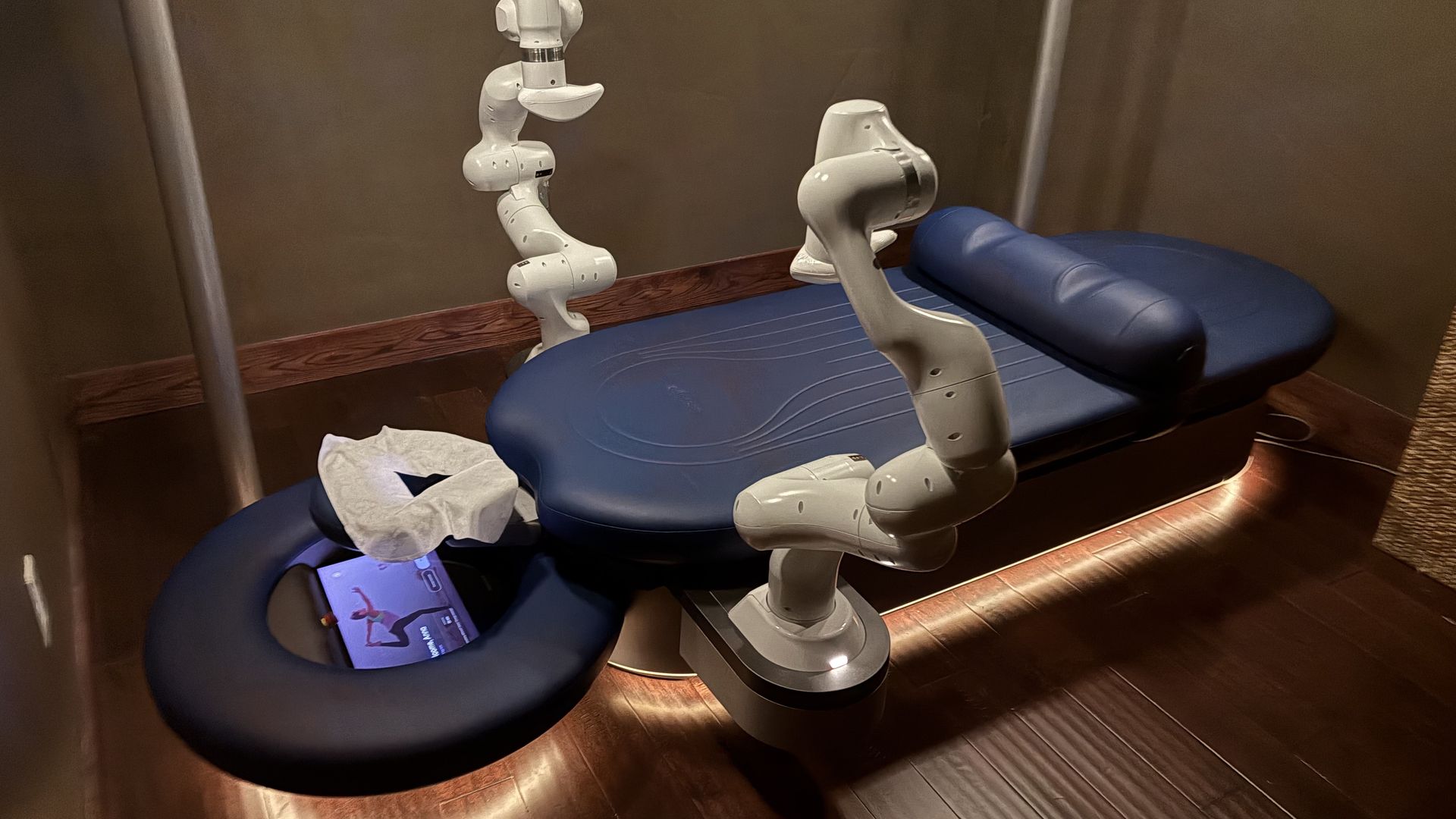 Dark blue massage table with white robotic arms in a dimly lit brown room with wooden floor and ambient lighting underneath the table.