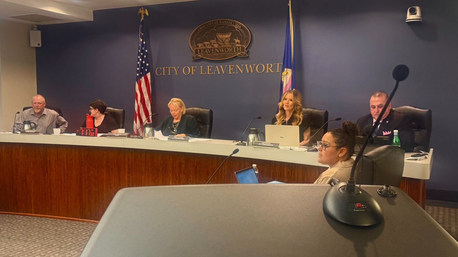 Five officials sit at a long desk in a city council chamber. A blue wall behind reads 'CITY OF LEAVENWORTH' with seal; flags, microphones, and laptops on the desk; a foreground attendee faces left.