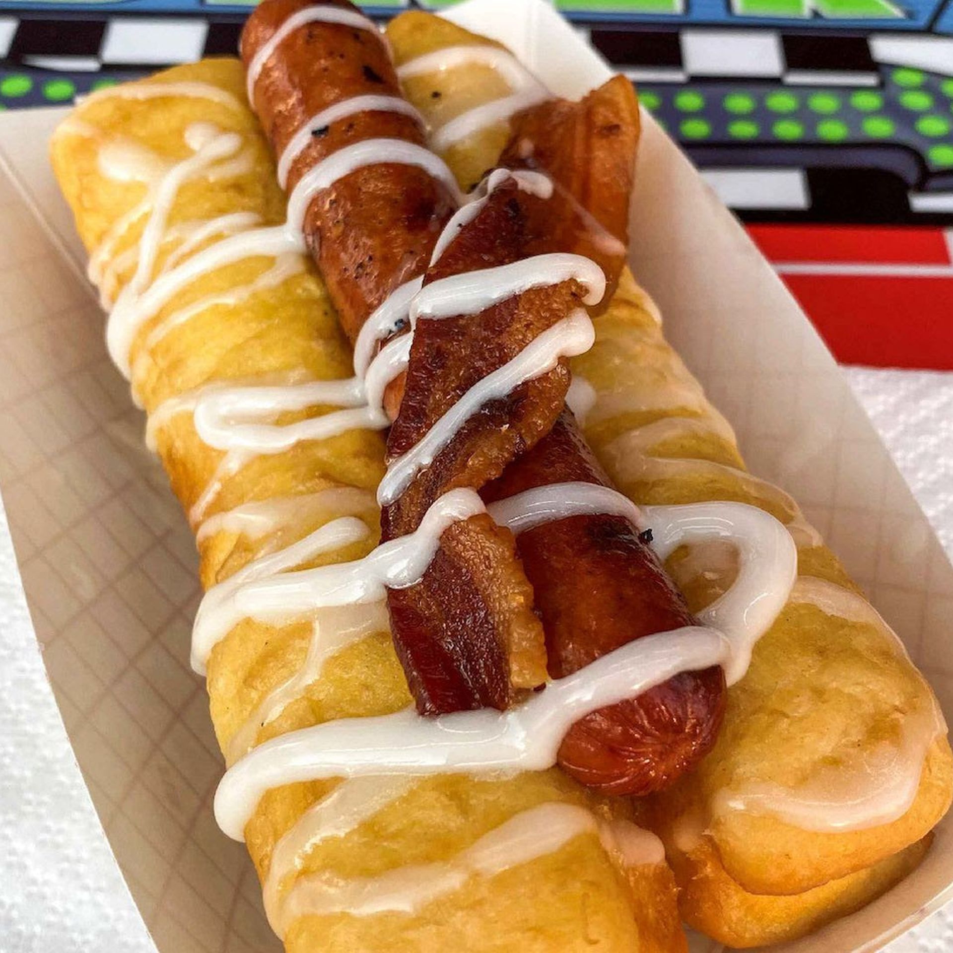  A homemade Long John donut wrapping a grilled all-beef hot dog topped with applewood bacon, all of it drizzled with icing on top.