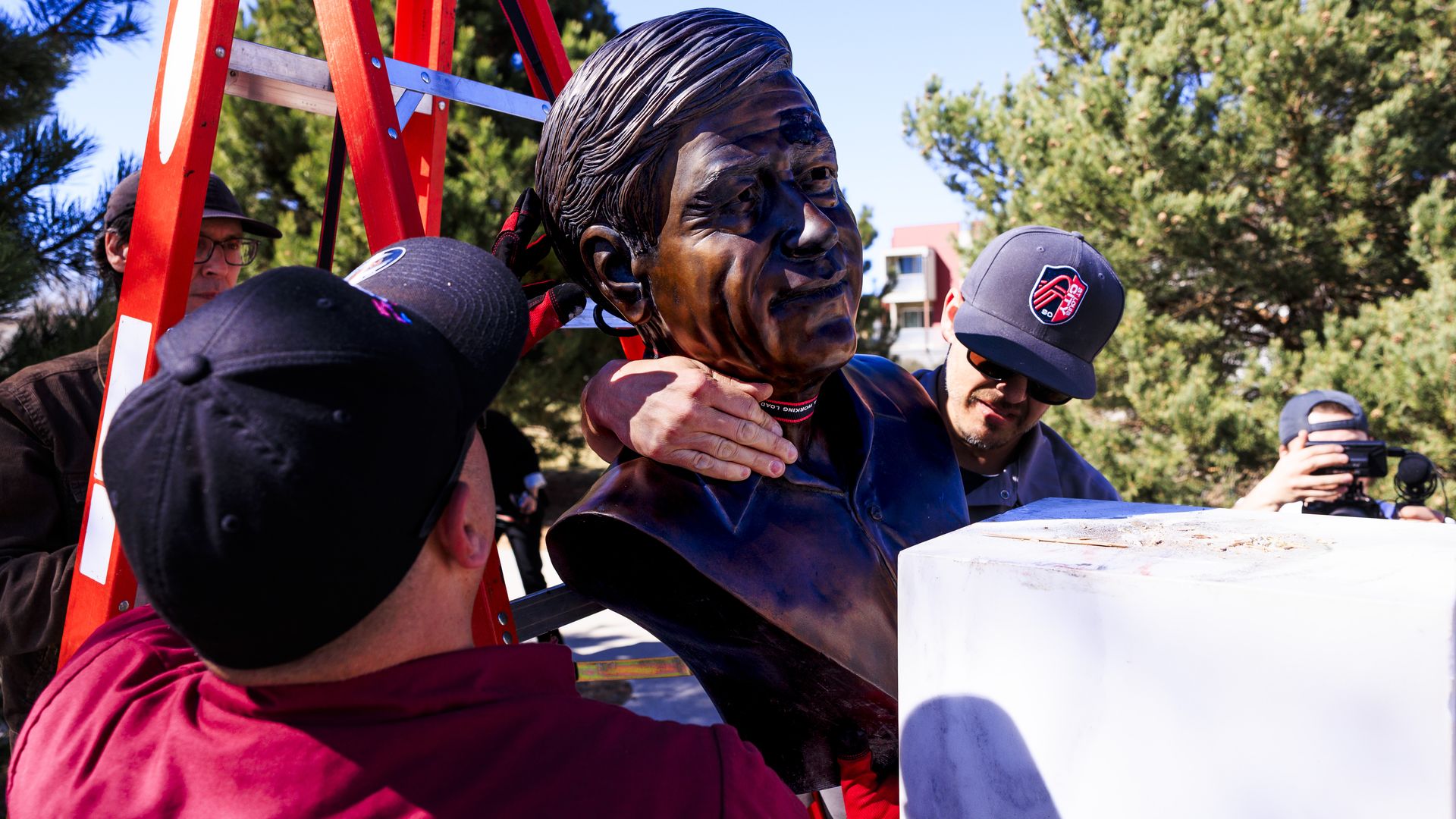 Outdoor scene of a bronze bust being gently adjusted by several people, with a red ladder, a white pedestal, and lush trees under a clear blue sky.