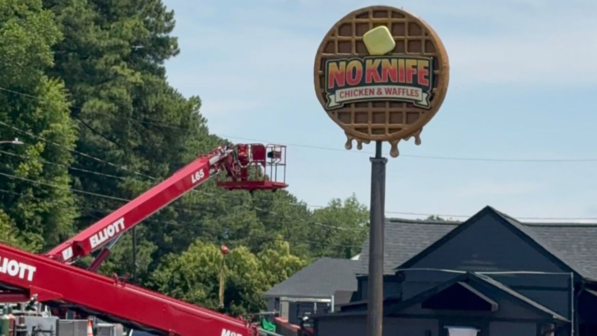 Giant waffle sign on a tall pole reads "NO KNIFE CHICKEN & WAFFLES" with a yellow square on top. Red Elliott construction lifts and workers in orange shirts line a street; trees and buildings in the background.