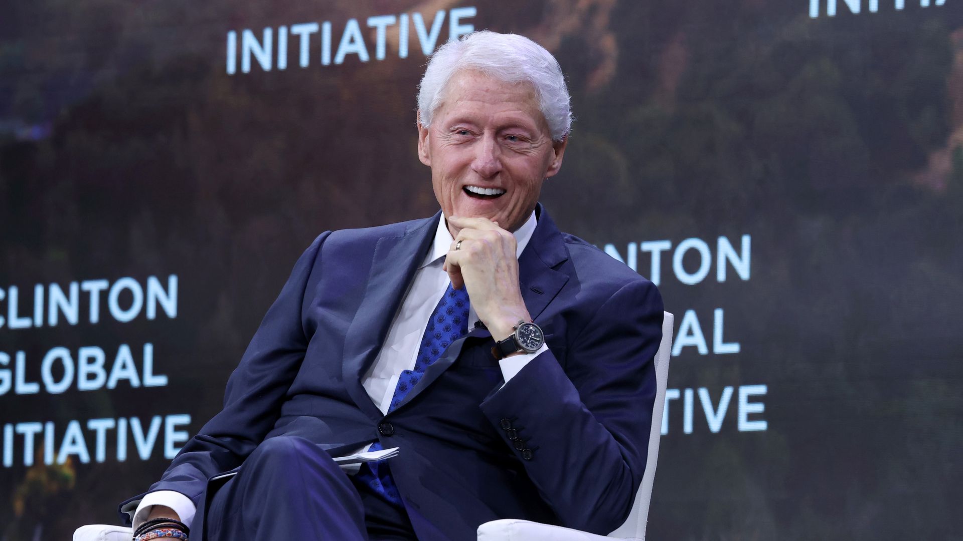 A man with white hair in a dark blue suit and blue patterned tie sits on a white chair, smiling and holding his chin, with a dark background showing "CLINTON GLOBAL INITIATIVE" text.