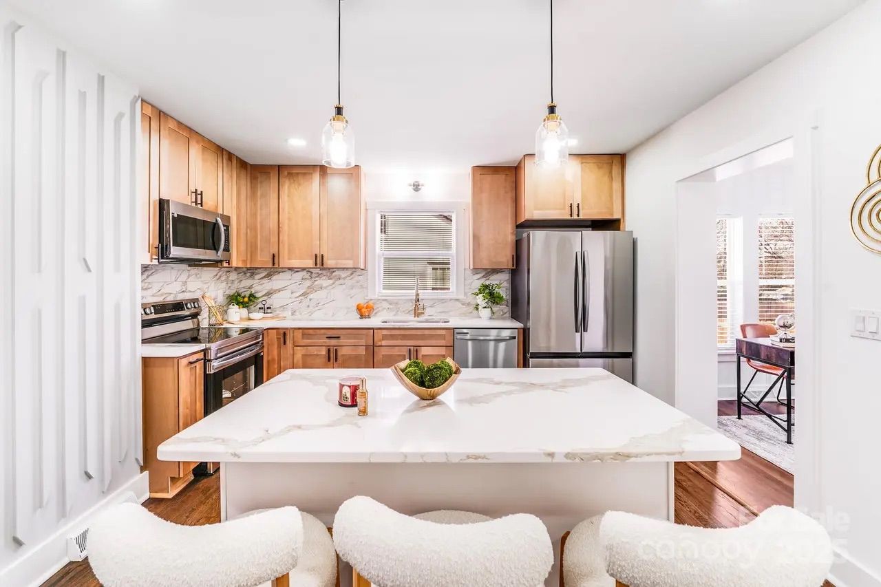 Bright modern kitchen with wooden cabinets, marble countertops, stainless steel appliances, an island with white cushioned stools, and pendant lights hanging from the ceiling.