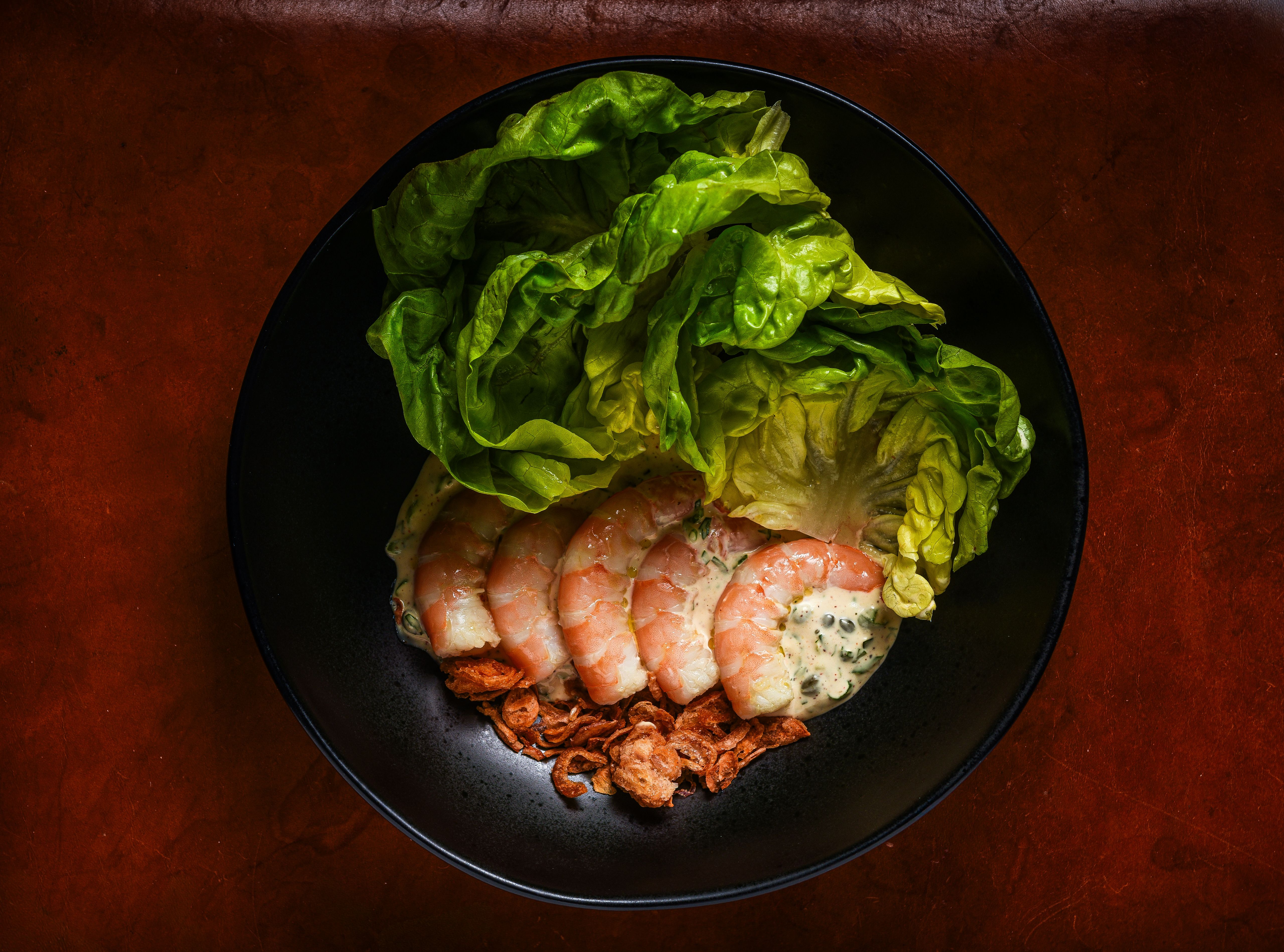 Black bowl with five cooked shrimp arranged over creamy sauce with herbs, topped with fresh green lettuce leaves and crispy fried shallots on a brown surface.