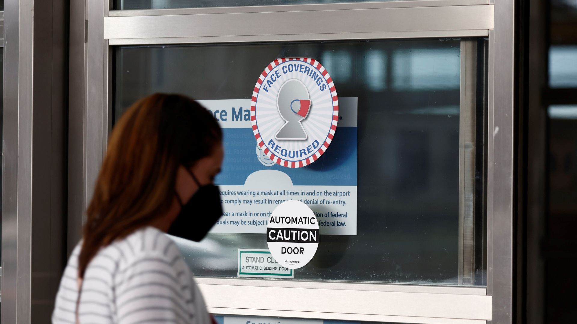 Photo of a masked person walking toward an automatic glass door that has a sticker saying "Face coverings required"