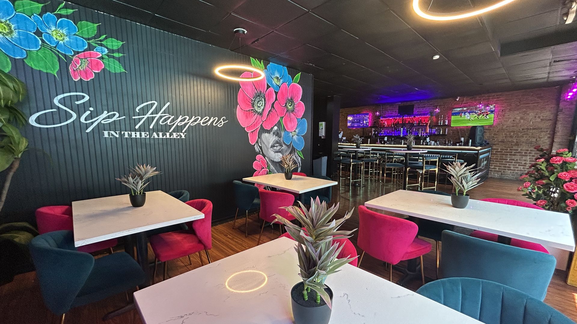 Trendy bar with a black mural of blue and pink flowers and a woman's face, "Sip Happens in the Alley". Neon ring lights, brick bar, and pink/teal seating around marble tables.