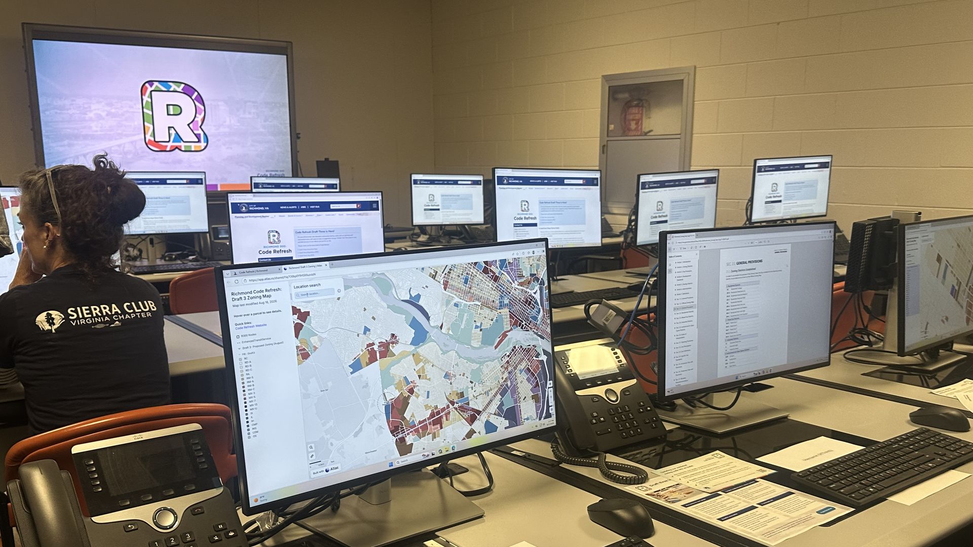 Row of computer workstations showing maps and web pages; a large screen on the left displays a colorful "R" logo. In the foreground a person wears a Sierra Club Virginia Chapter shirt.