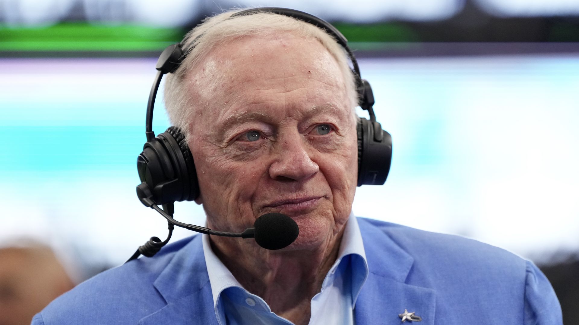 A person in a light-blue suit and collared shirt speaks into a headset microphone