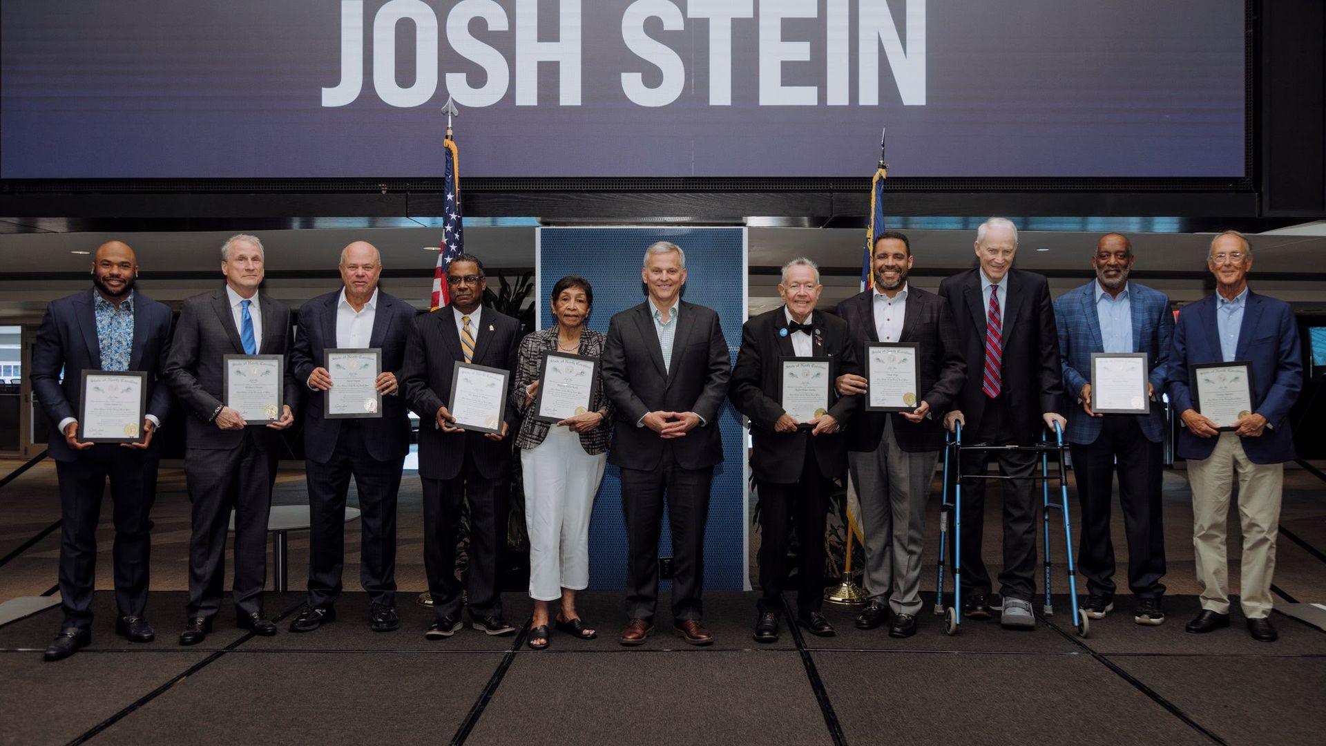 Line of diverse award recipients in suits, holding certificates on a stage, flanked by two flags, with a large screen displaying the name "JOSH STEIN" behind them.