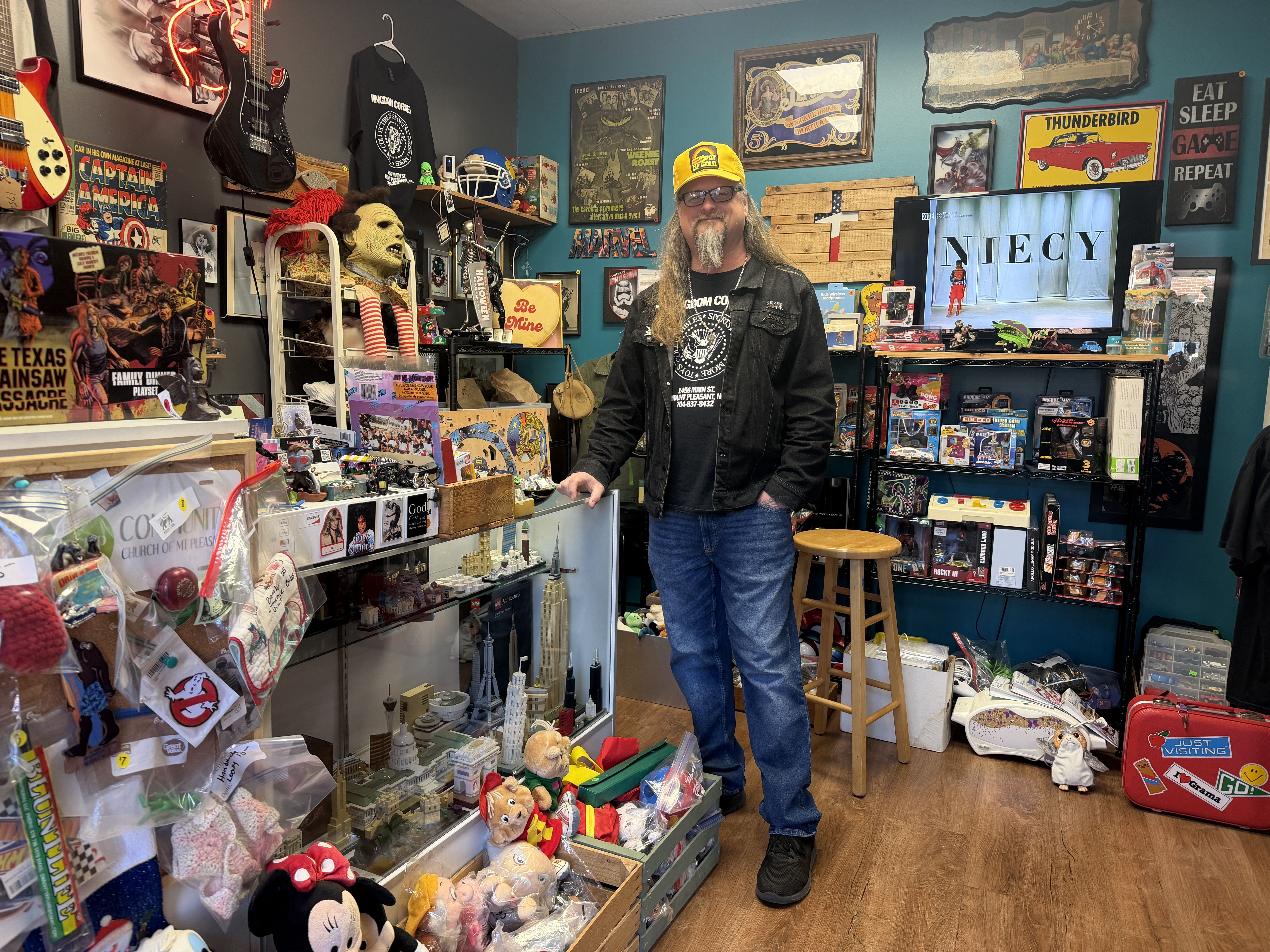Man with long hair, yellow hat, black jacket and jeans stands in eclectic room filled with collectibles, toys, vintage posters, model buildings, and signs including "Eat Sleep Game Repeat" and "Thunderbird."