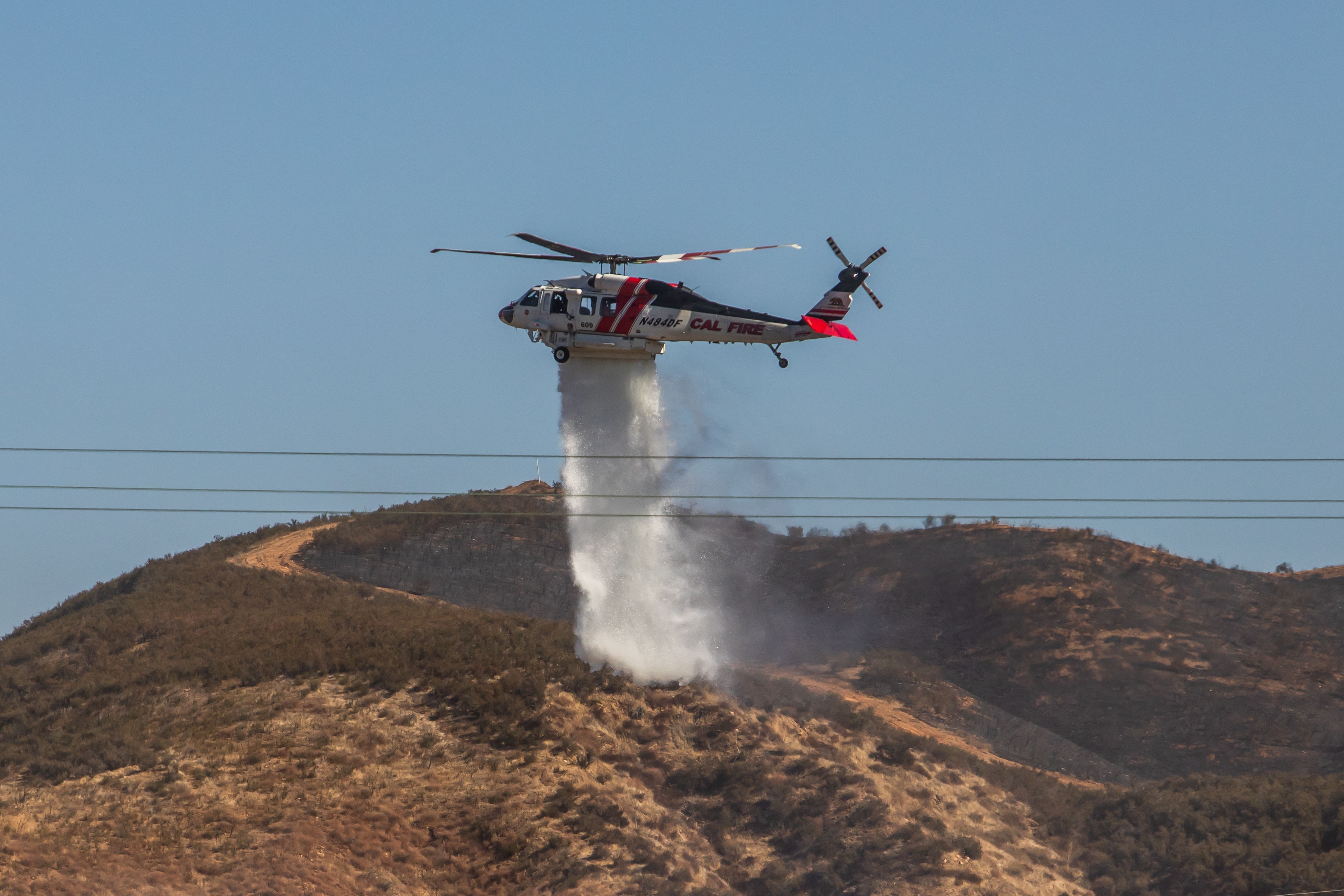 A CAL FIRE helicopter hovers above a dry, brush-covered hillside, releasing a large column of water onto the terrain below. 