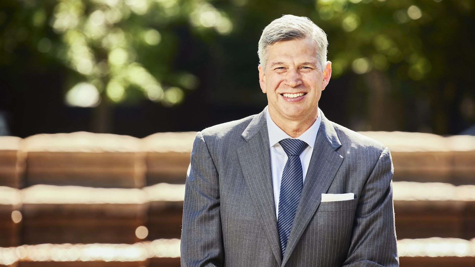 A photo of a person wearing a gray two-piece suit smiling at the camera in front of a blurred background.