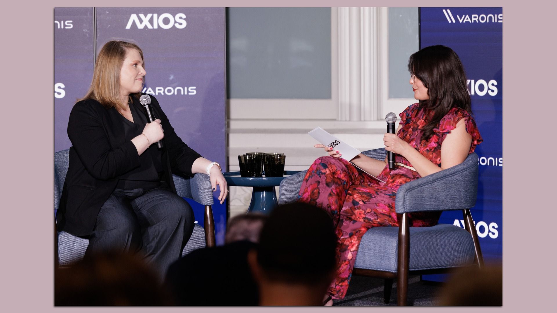 Two women sit on stage in blue chairs during a talk-show interview. Left: blonde in a black blazer; right: woman in a red floral dress holding notes. A small table with drinks and an Axios backdrop.