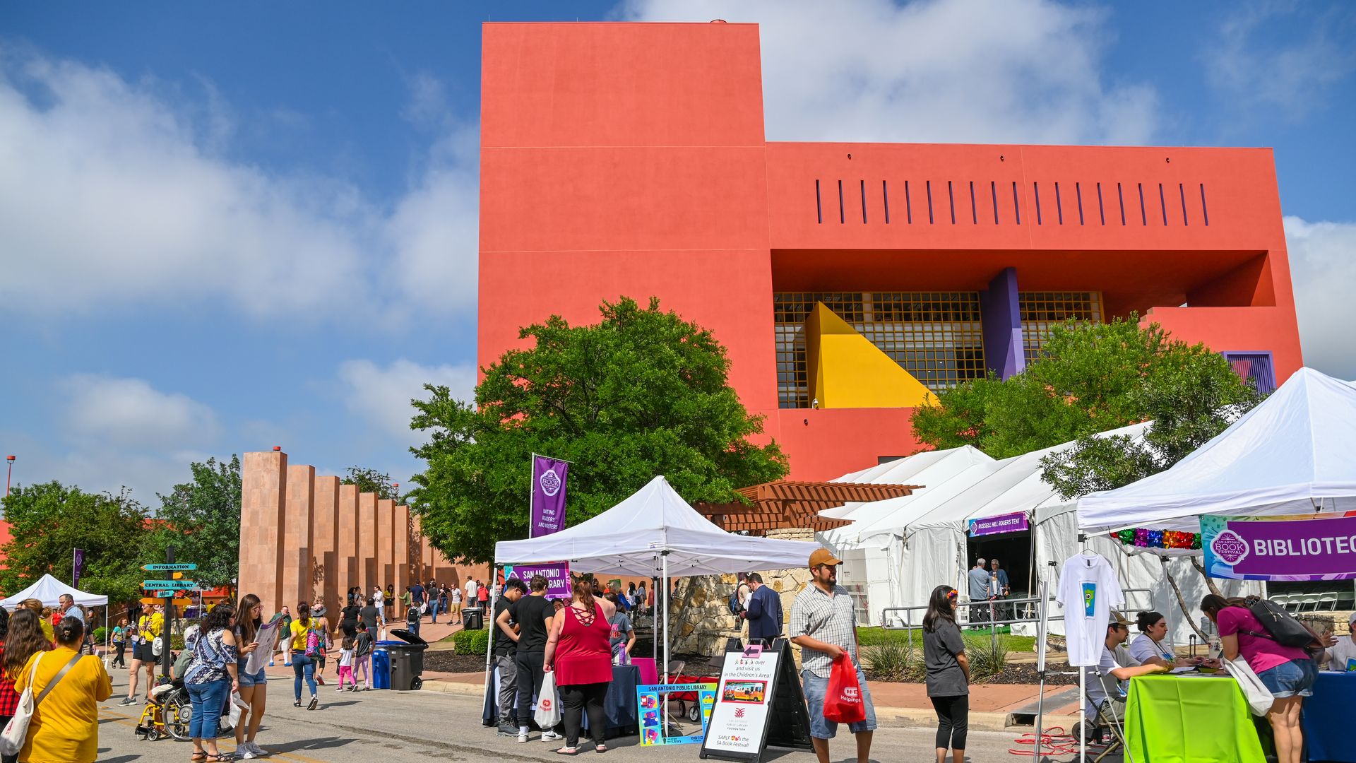 People gather under white tents near a orangey, red and modular building. 