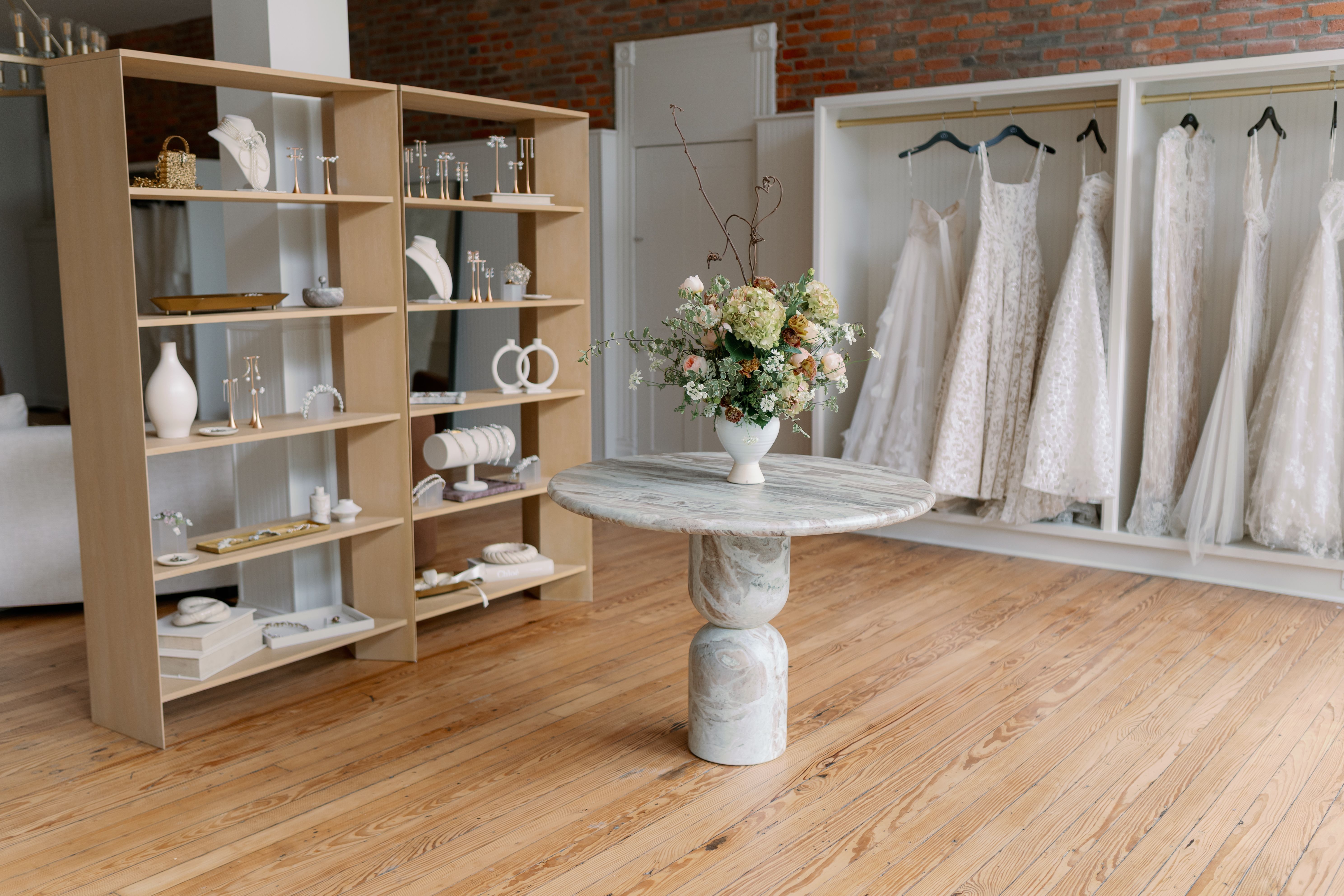Bridal boutique interior with wooden floor, marble round table holding a floral vase, wooden shelves displaying jewelry, and white wedding dresses hanging on racks against a brick wall.
