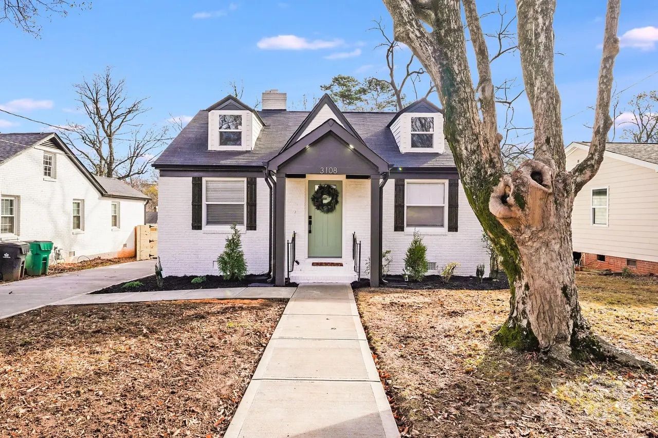 White brick house with black trim and a green front door decorated with a wreath. A sidewalk leads to the door. Large tree with thick branches stands to the right in a leaf-covered yard.