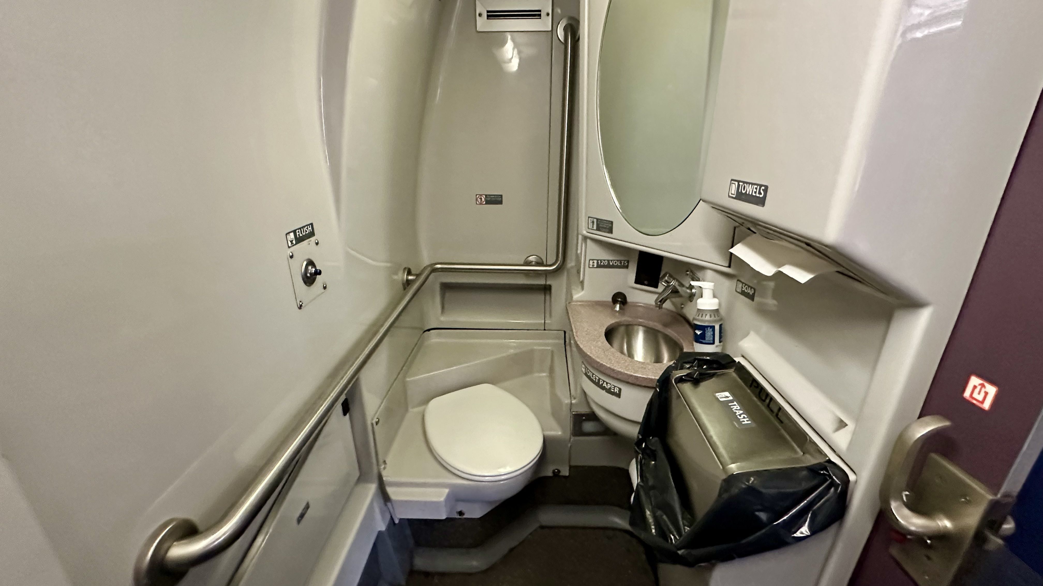 Compact airplane lavatory with white toilet, sink with pink countertop, wall-mounted soap, towels, and trash bin, metal handrails, and safety labels including a flush sign.