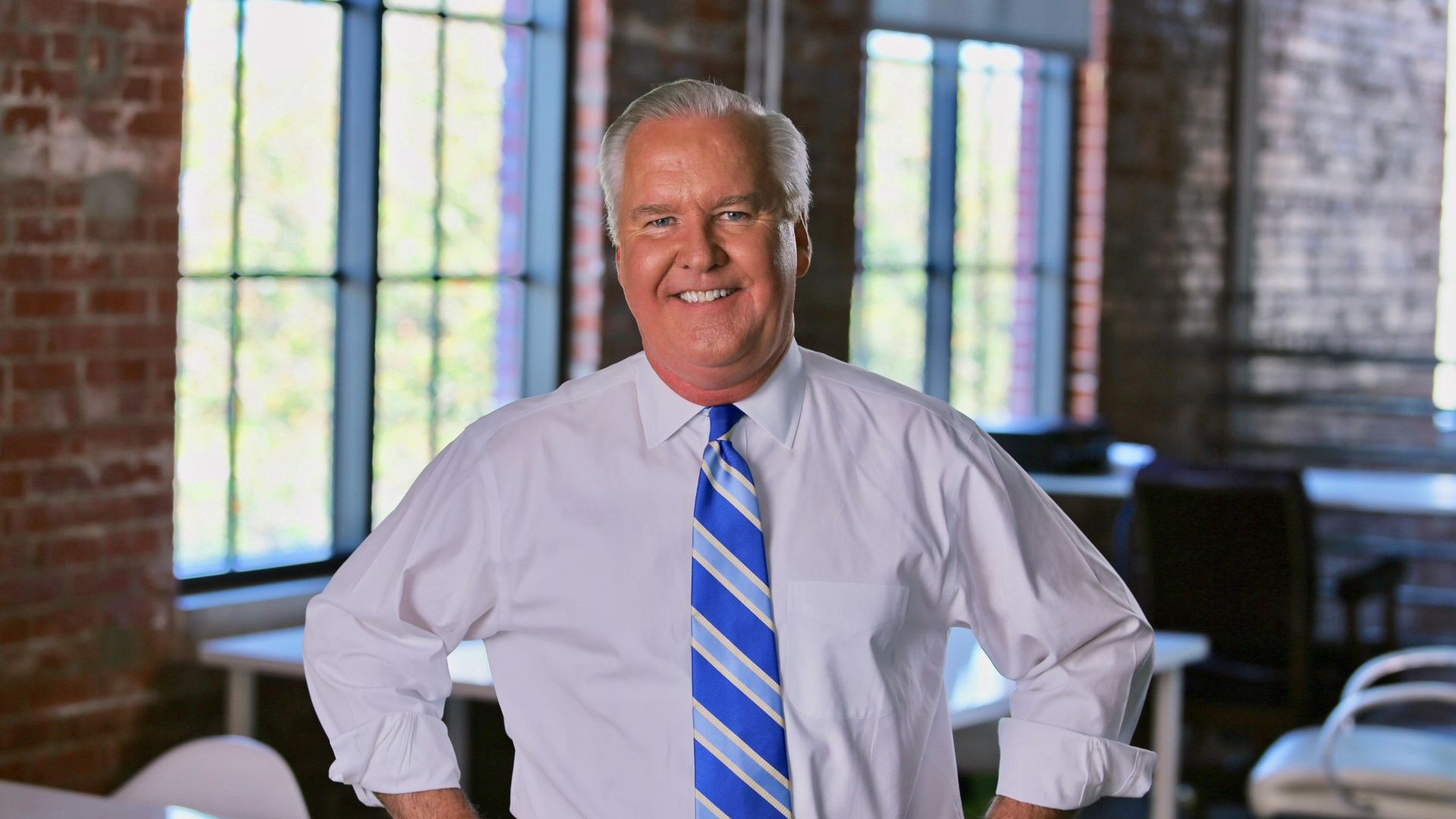 Smiling older man with white hair in a white dress shirt and blue striped tie, standing with hands on hips in a bright brick-walled room with large windows.