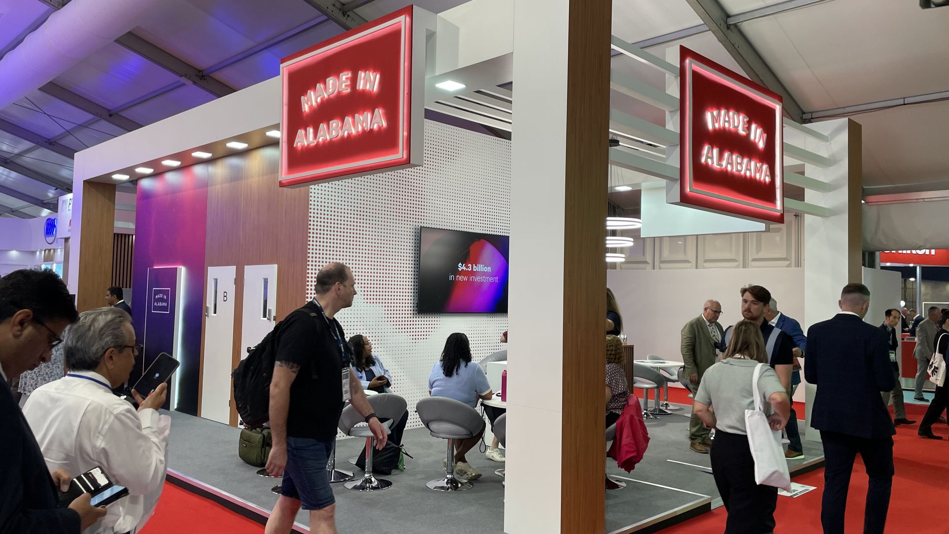 Trade show booth with white and wood panels, bright red neon signs reading "MADE IN ALABAMA," red carpet, and groups of people walking and talking around the exhibit.