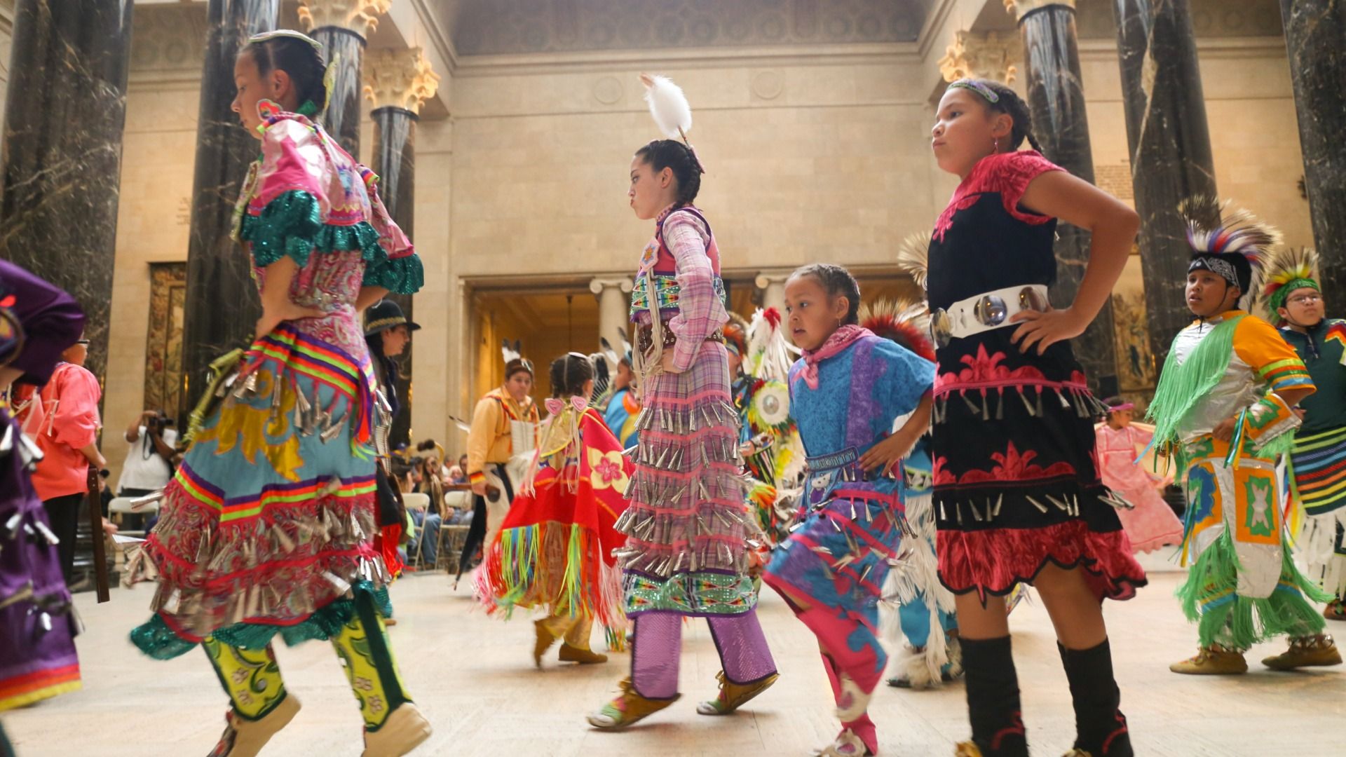 Group of children in colorful traditional Native American regalia dancing indoors in a hall with tall dark marble columns and beige walls.
