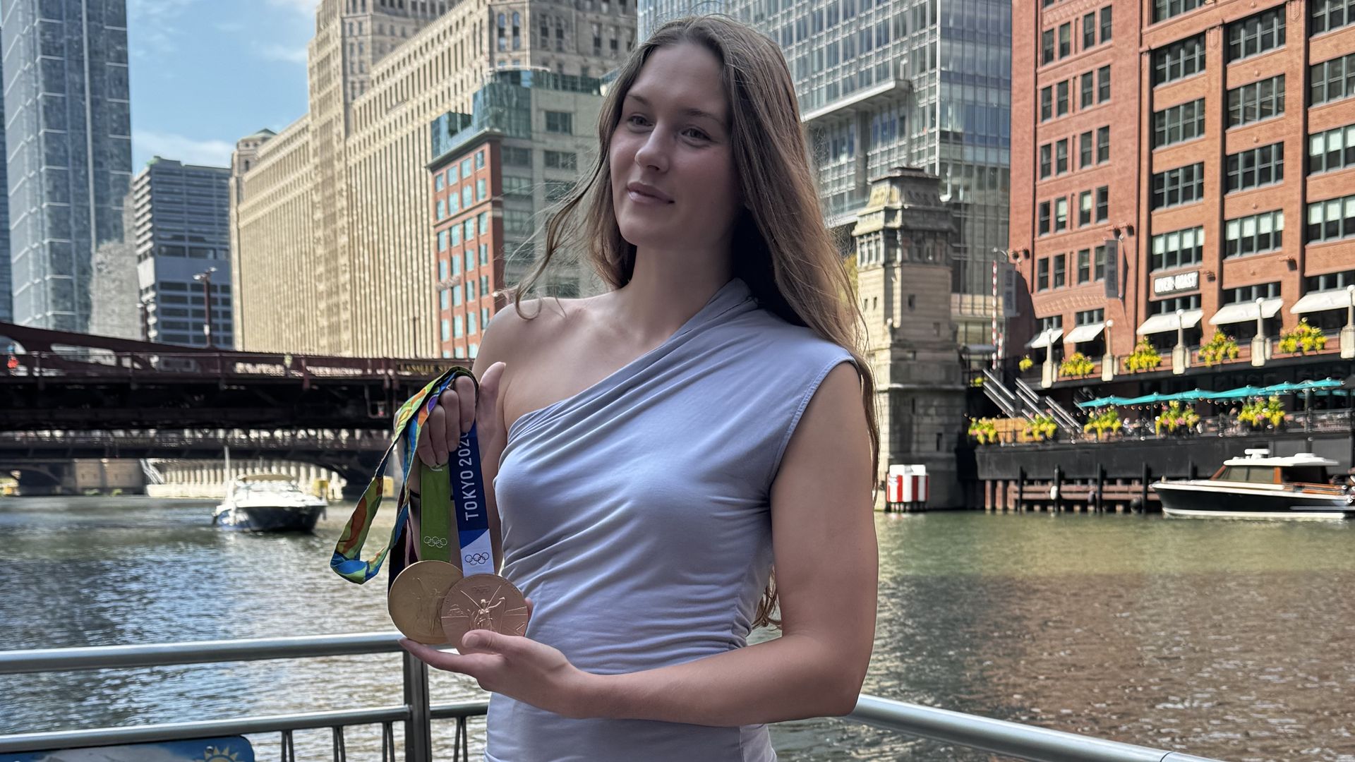 Young woman with long brown hair wearing a light blue one-shoulder top stands by a railing along a river, holding Olympic medals; city skyline with tall buildings, a bridge, and boats behind her.
