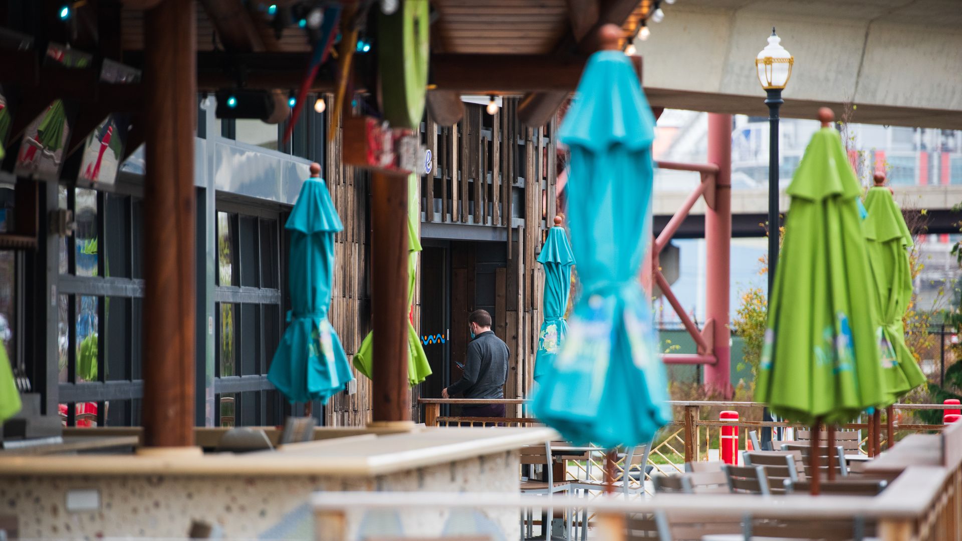 Outdoor restaurant patio with closed green and blue umbrellas, wooden tables and chairs, and a person in dark clothing using a phone in the background near a wooden building.