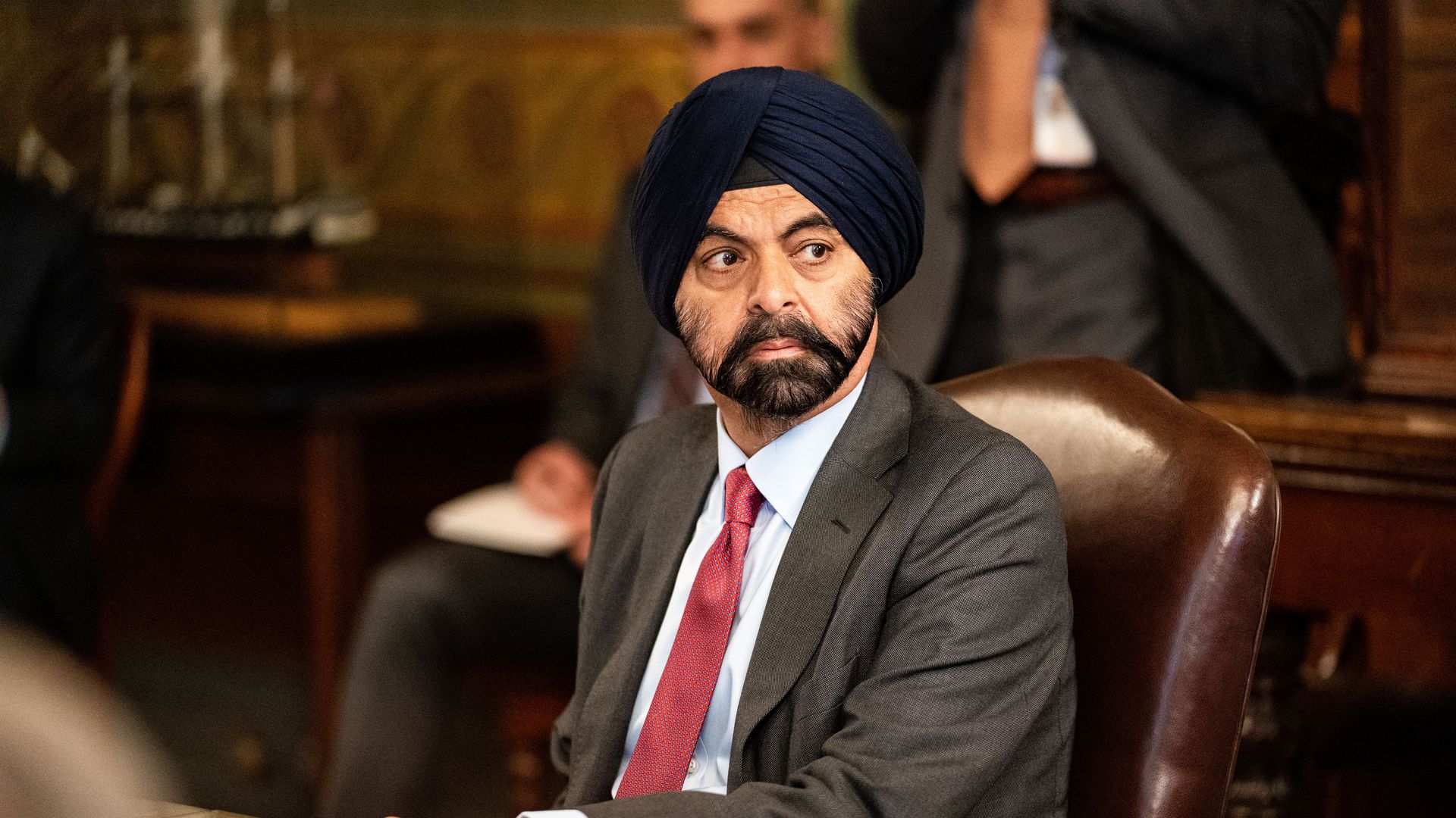 A bearded man in a gray suit, red tie and Sikh turban looks to his left as he is seated in a rich burgundy leather chair
