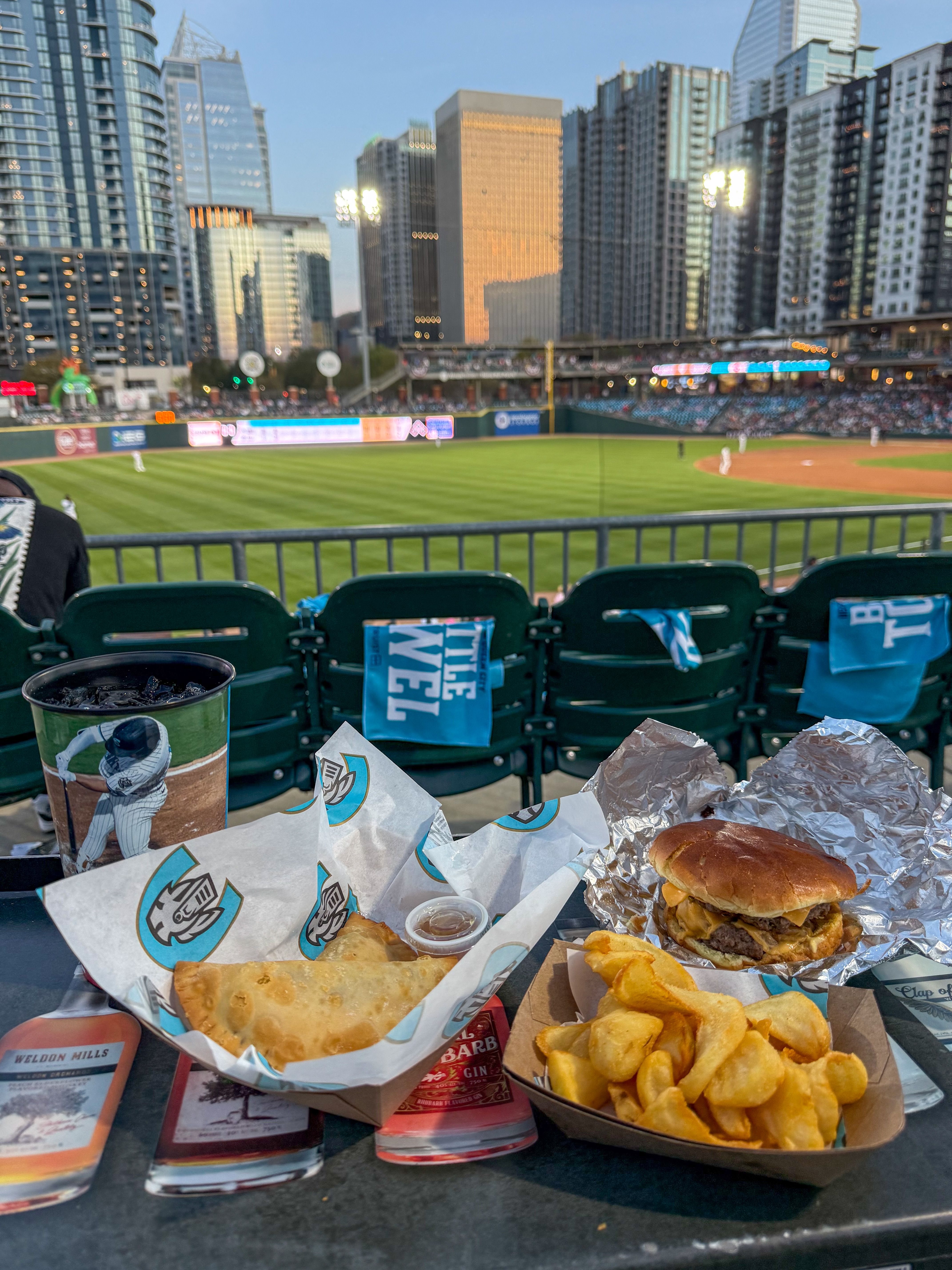 A tray with empanadas, curly fries, a cheeseburger and a drink is set on a counter overlooking the outfield.