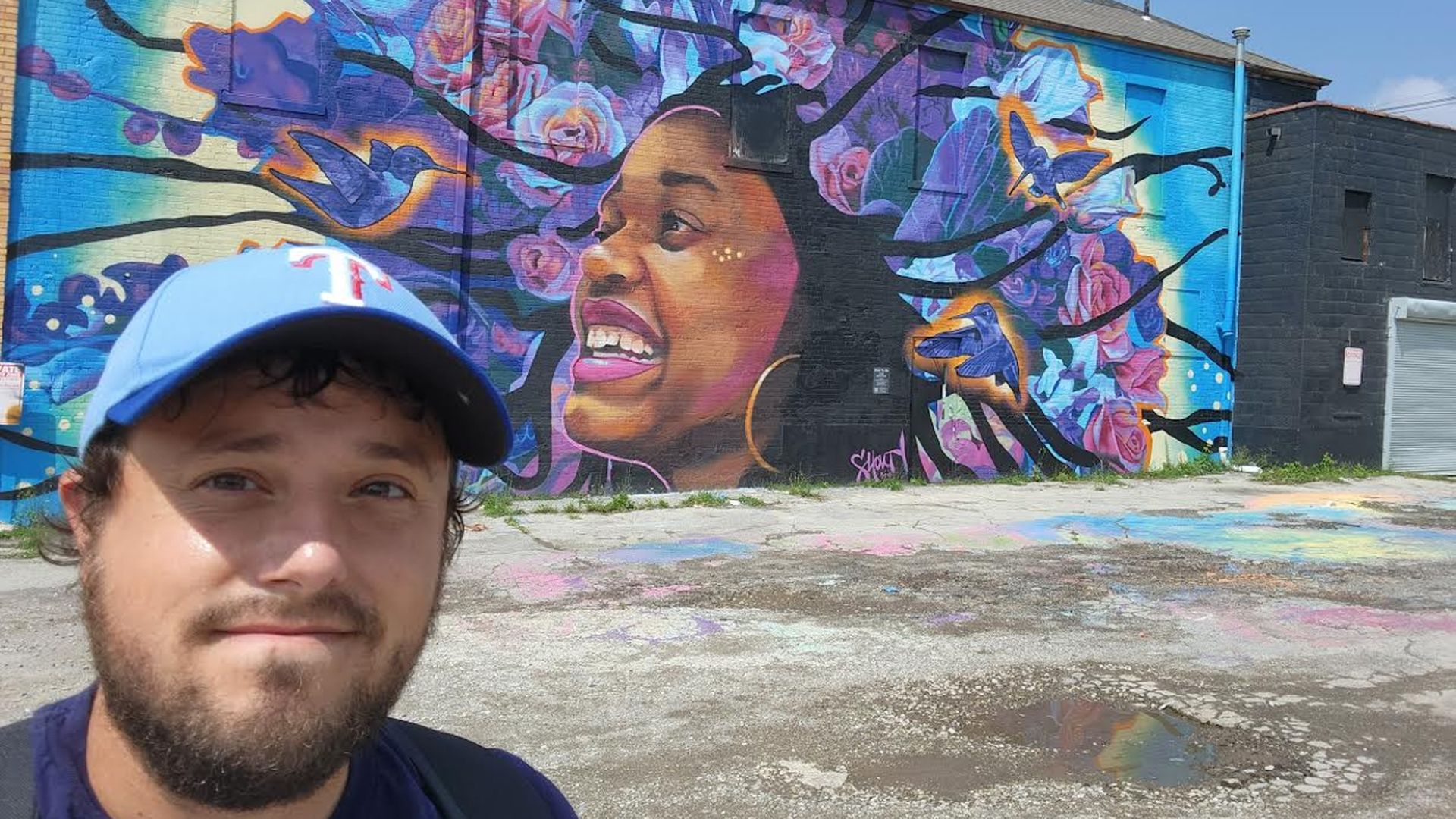 Tyler takes a selfie in front of a mural of a woman's head surrounded by butterflies, birds and flowers