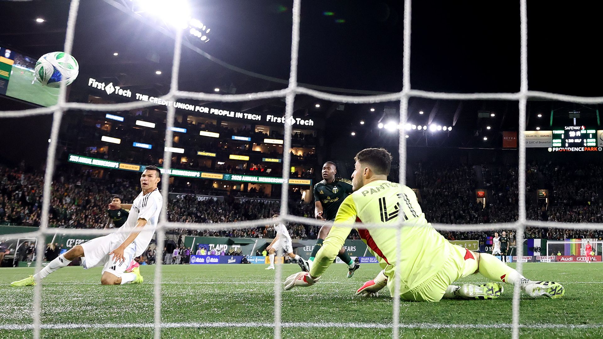 A view from behind the net of a soccer goal of San Diego FC player Chucky Lozano sliding to score past a goalkeeper in a neon jersey. 