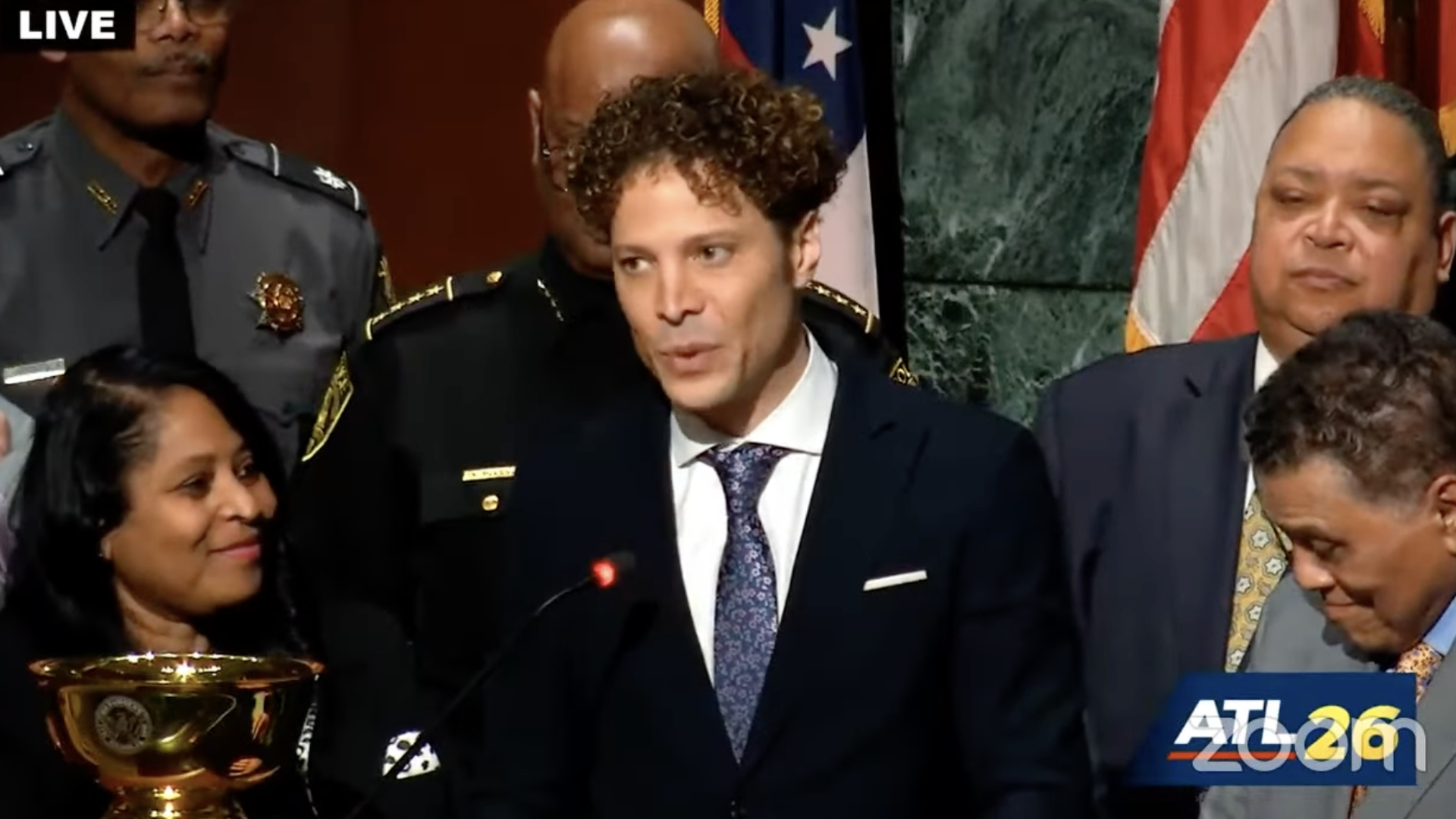A young man with curly hair in a dark suit stands at a microphone to pay tribute to another person. A gold cup sits on the lectern.