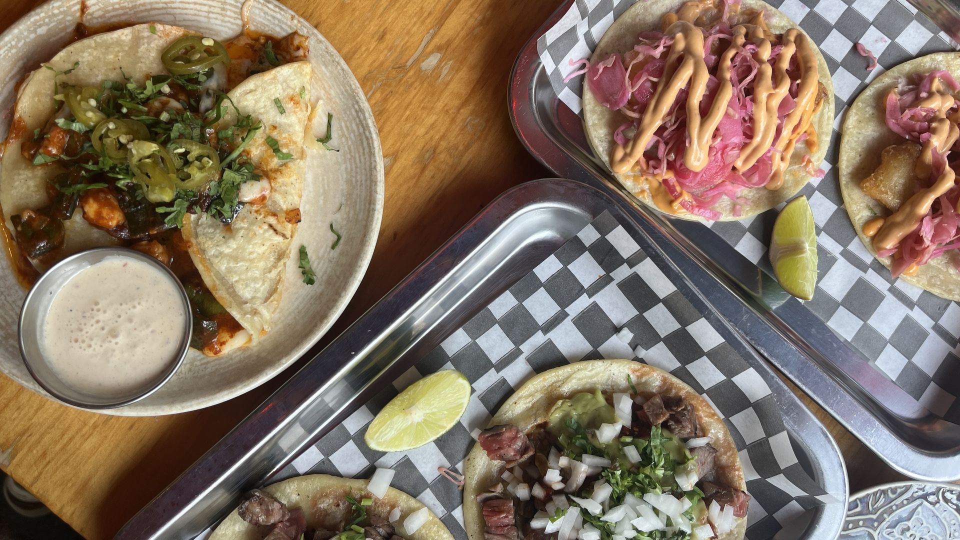 Overhead view of tacos on a wooden table: left plate with cilantro and jalapeños; right tray with pink pickled onions and orange crema, lime wedges, and checkered parchment.