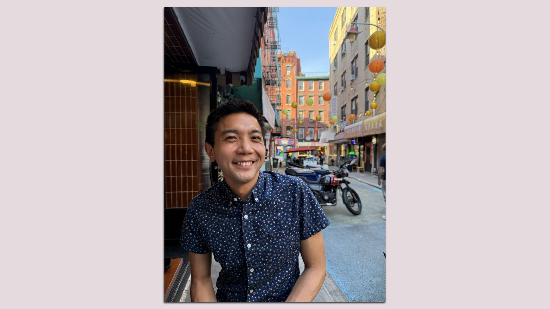 Smiling man in a navy blue floral shirt sits at a sidewalk cafe on a busy city street, with colorful lanterns overhead, brick buildings along the road, and a motorcycle parked nearby.