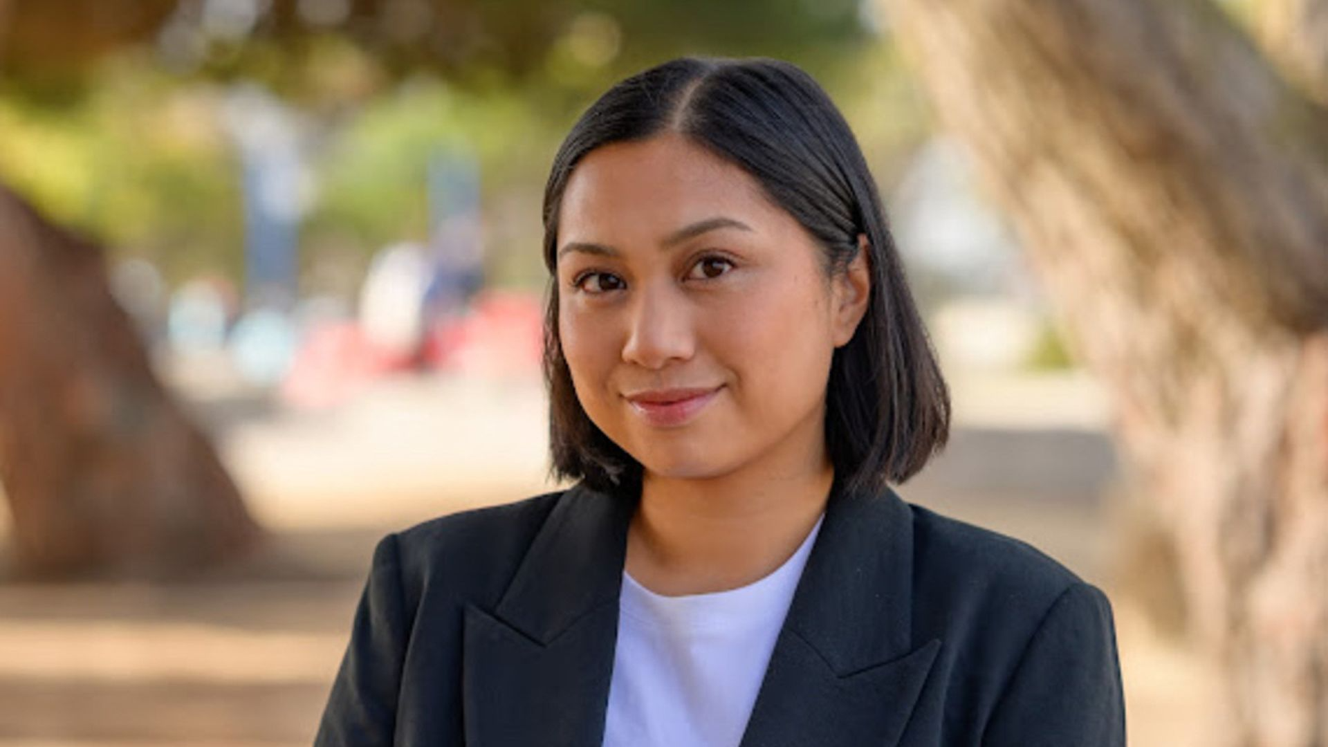 Smiling woman with short black hair wearing a black blazer and white shirt, outdoors with blurred trees and people in the background.