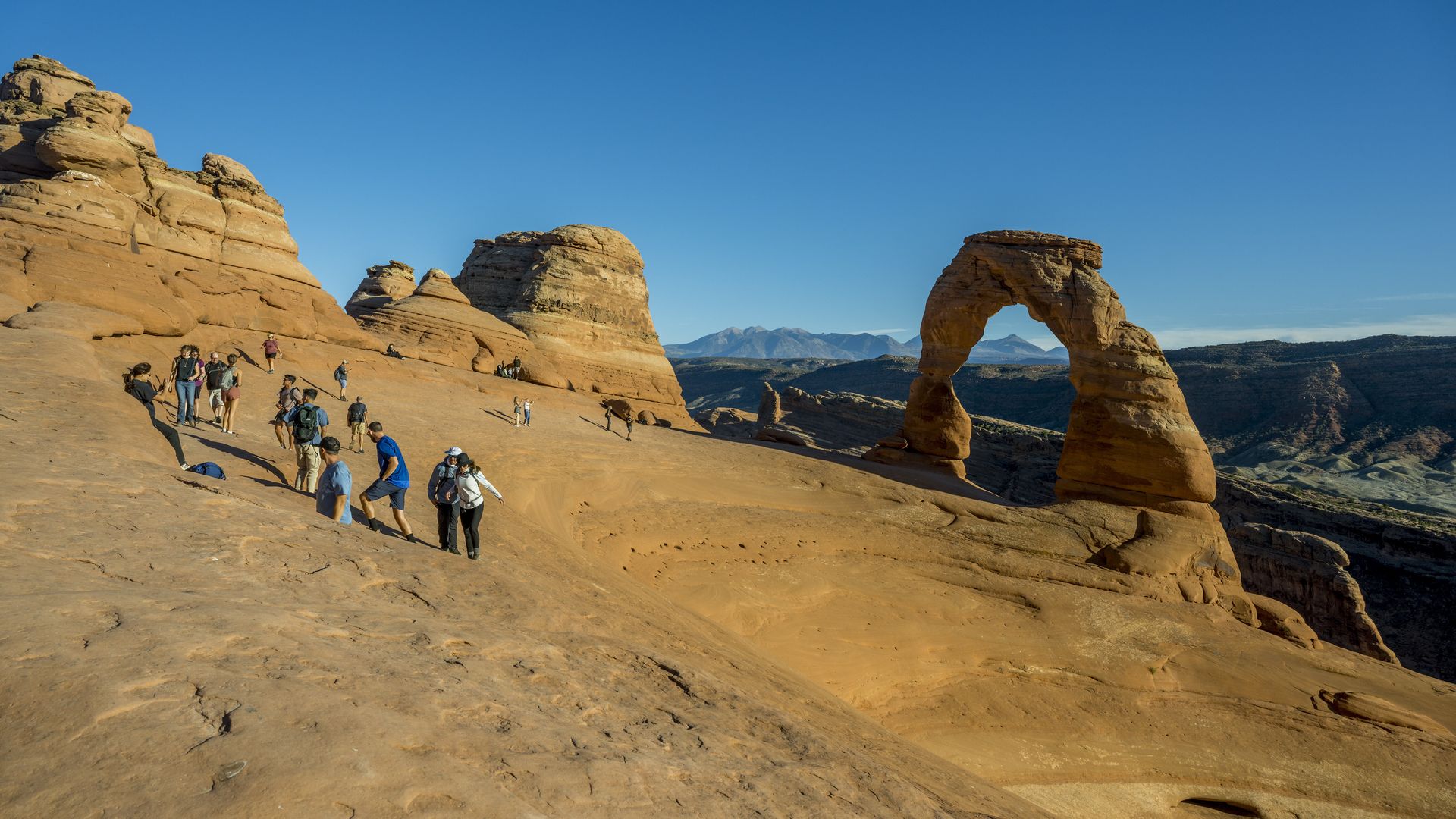 Desert scene with orange sandstone formations and a large natural arch under a clear blue sky. A group of hikers with backpacks traverse the sandy ground, distant mountains on the horizon.