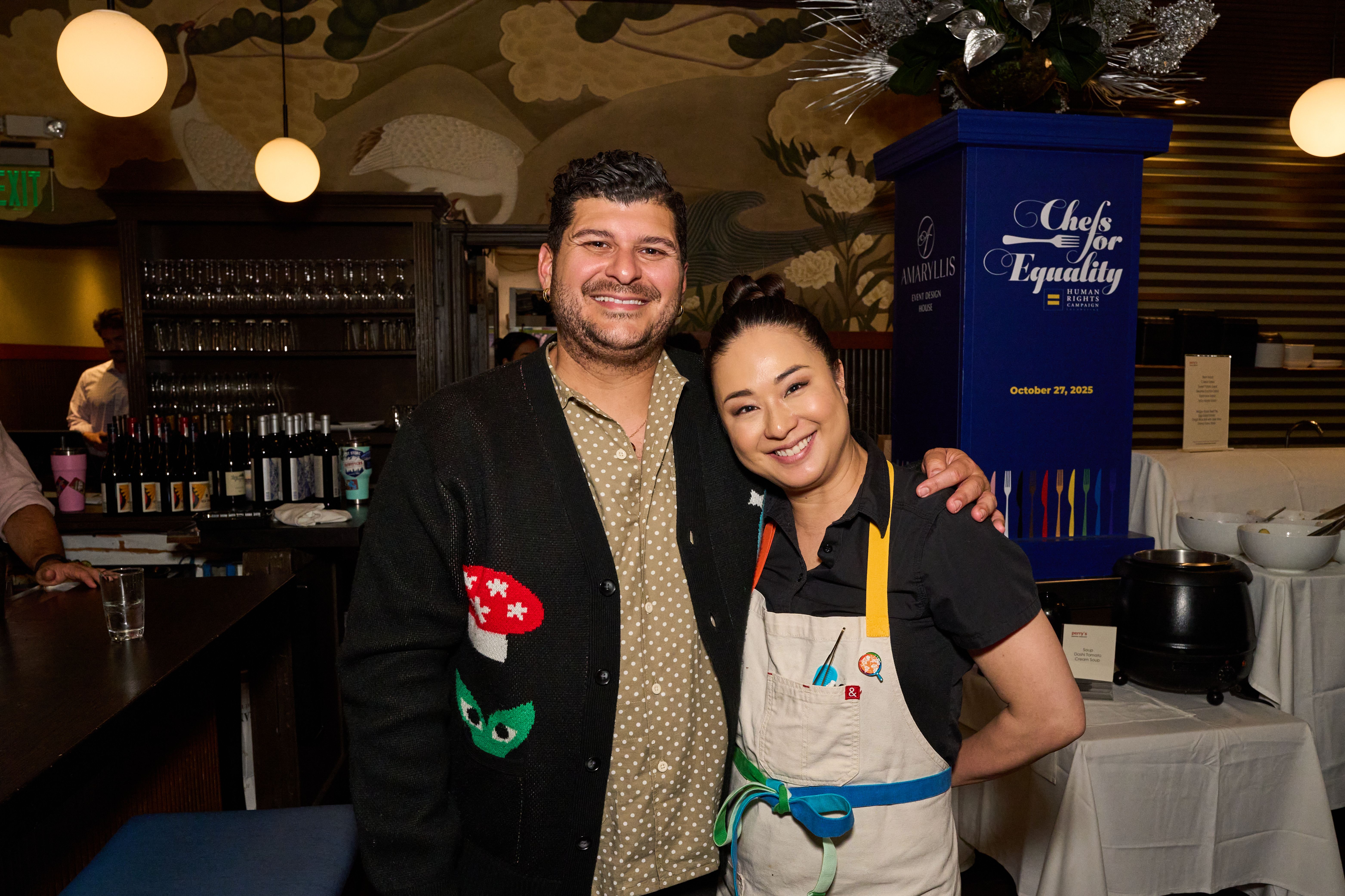 Two smiling people pose at a bar: a man in a beige polka-dot shirt and dark cardigan, and a woman in a white apron with blue straps. Behind them a blue pillar reads "Chefs for Equality" beside glassware shelves.