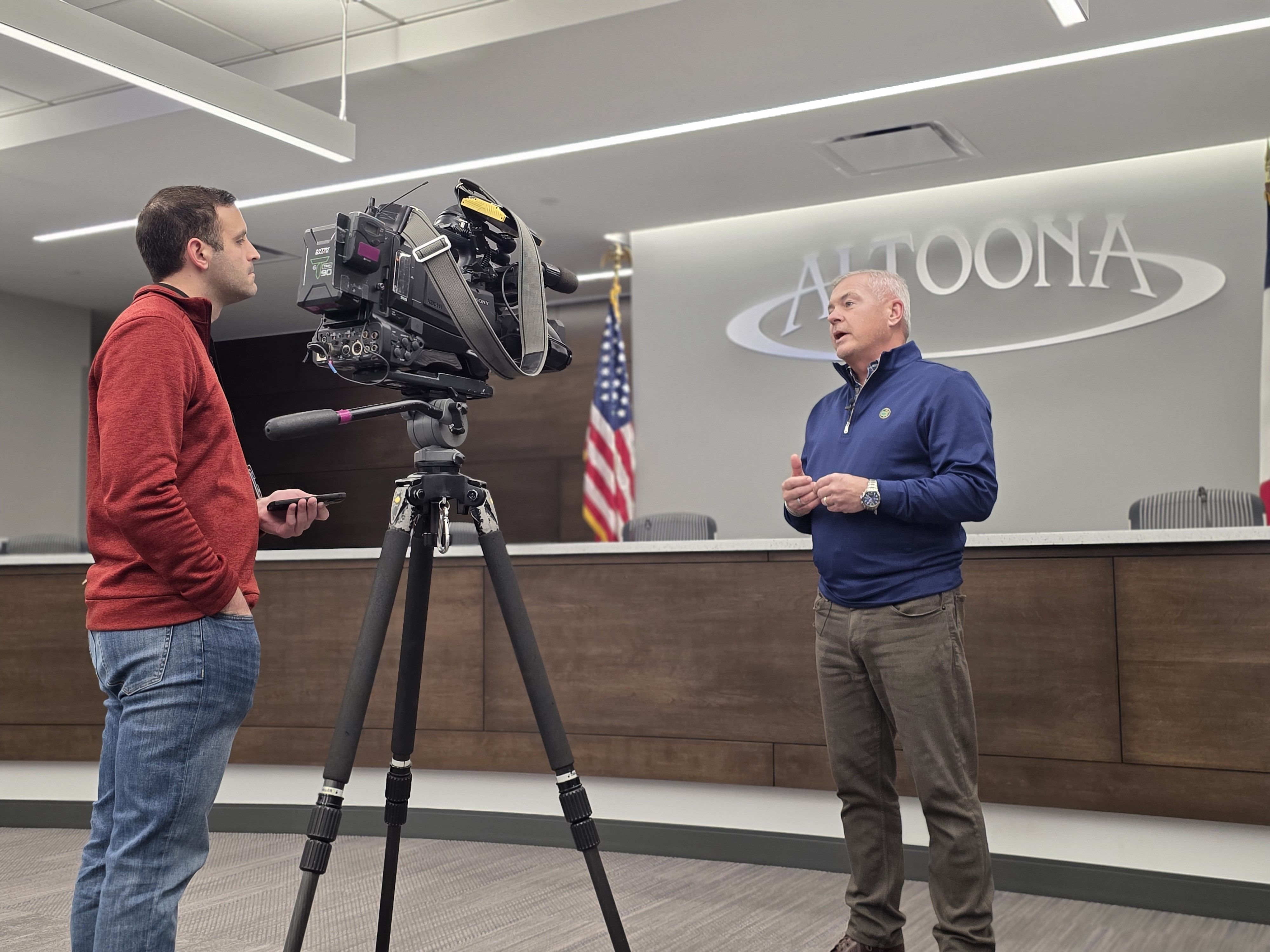 Man in red sweater operates video camera on tripod filming another man in blue pullover speaking, standing in council chamber with American flag and "ALTOONA" sign on wall.