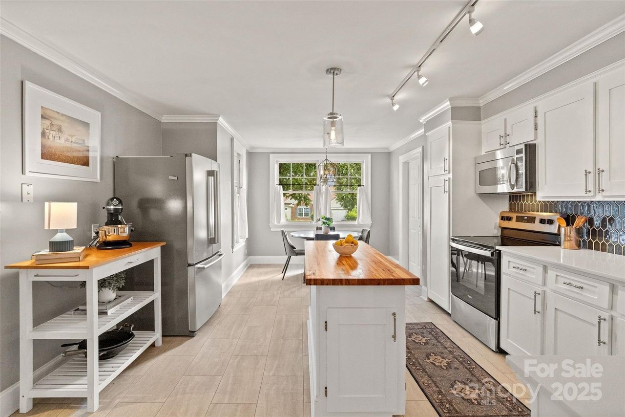 Bright kitchen with white cabinets, wooden countertop island, stainless steel fridge and oven, gray tiled floor, blue tile backsplash, small dining table near window, and decorative lamp.