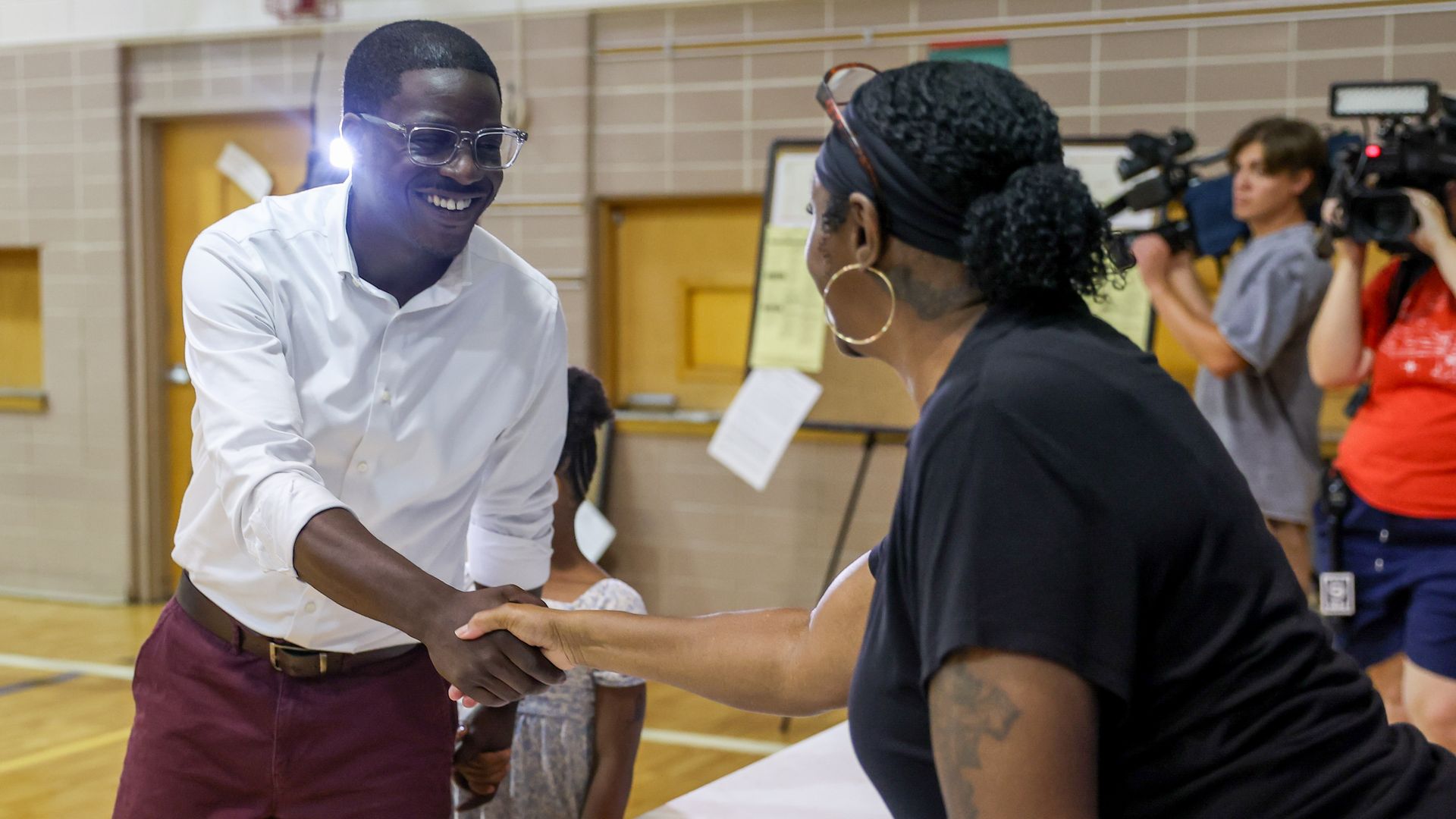 Crowley, wearing a white shirt and glasses, greets a woman inside a Milwaukee polling place as news cameras record the interaction.