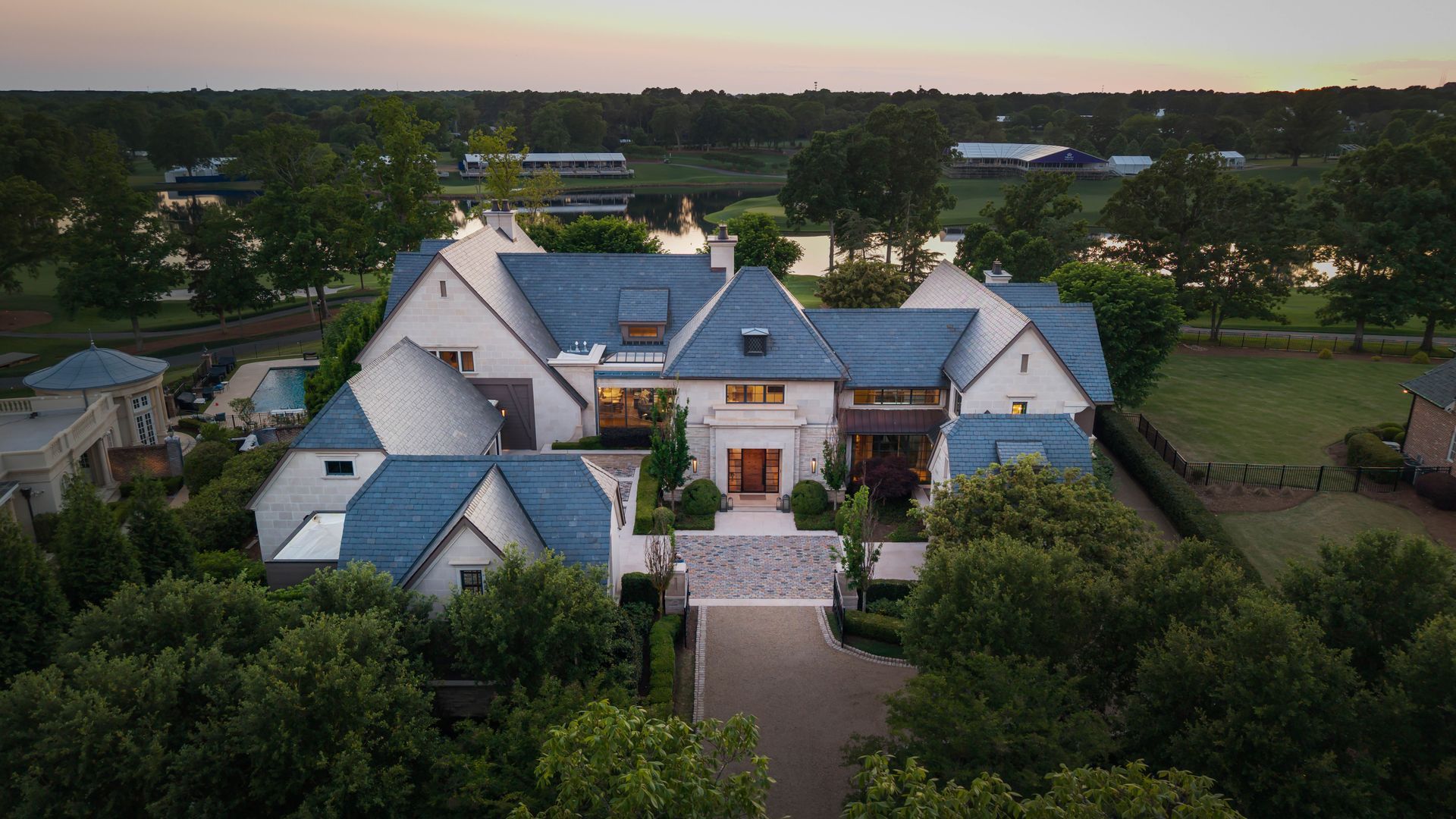 An aerial view of a grand stone house with blue slate roofs, a curved driveway, and manicured hedges. Surrounded by trees, with a lake and sunset sky in the distance.