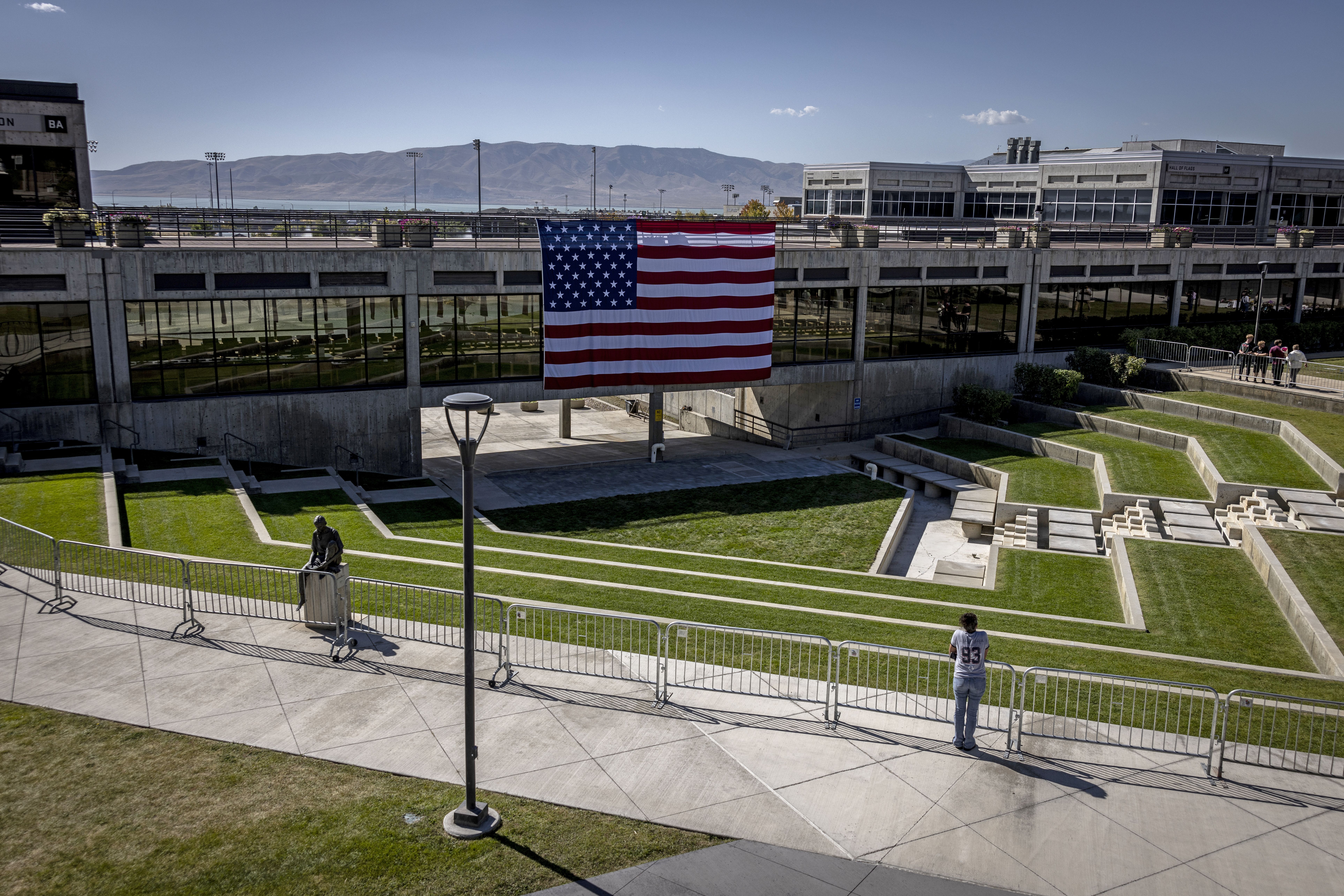 Large American flag hanging on a modern concrete building with glass windows, terraced green lawn, barriers, a lone person, and distant mountains under a clear blue sky.